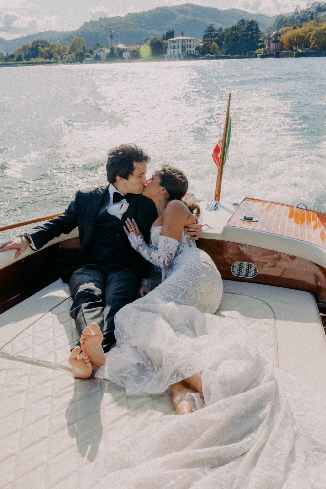 A romantic couple lounges together on a boat deck with scenic mountain and lake views in the background.