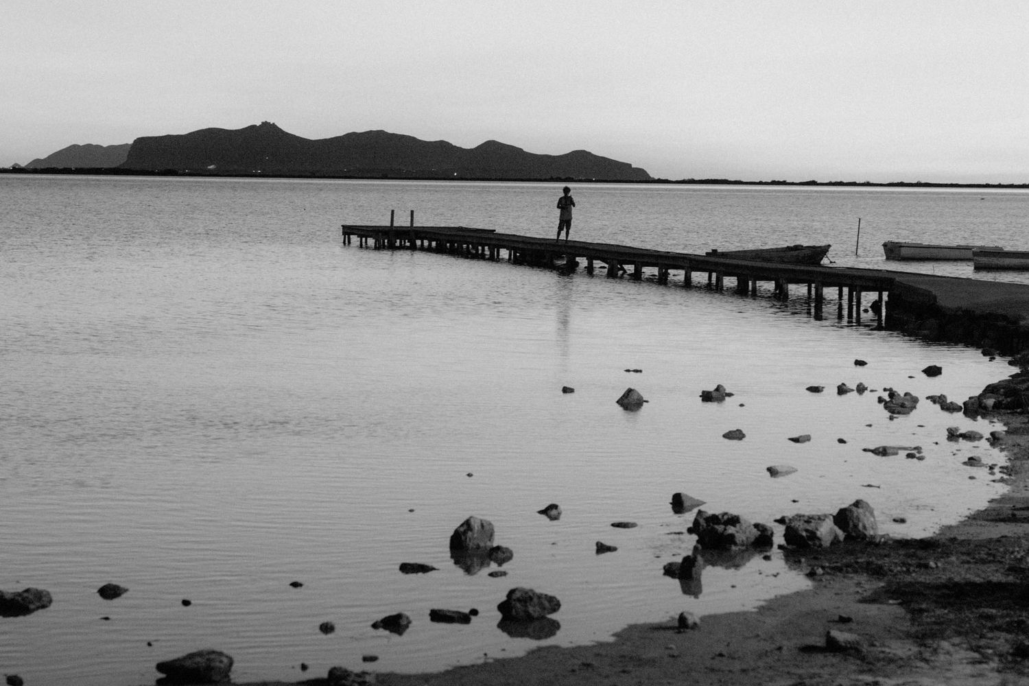 Silhouetted pier extends into calm water with mountains in background and scattered rocks along shoreline.