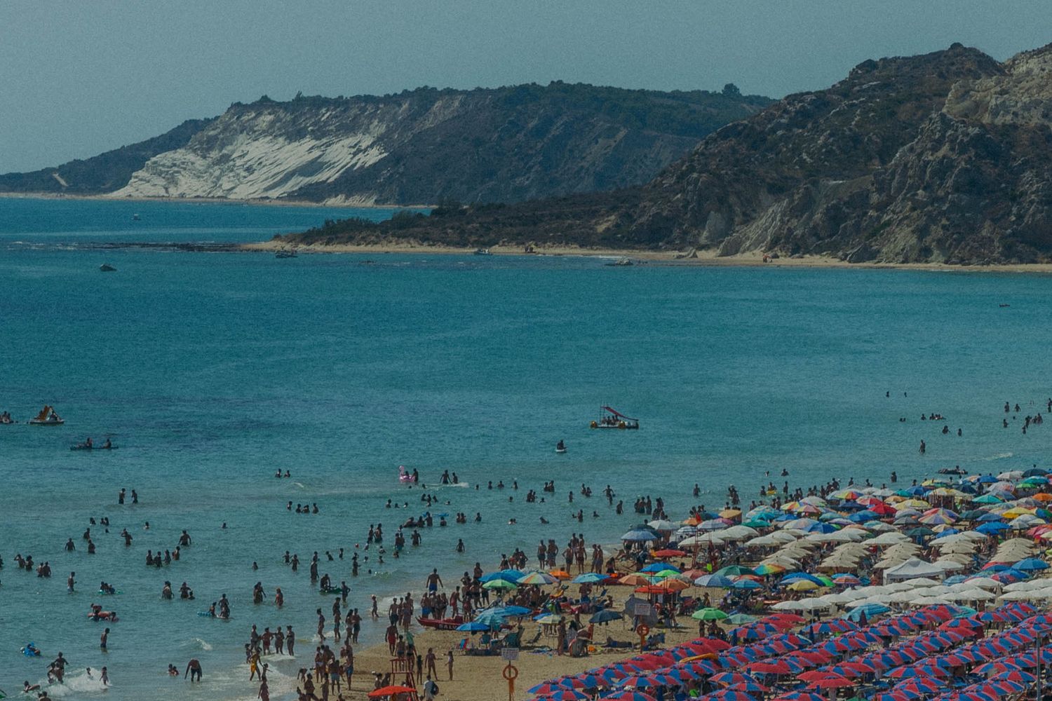 Crowded summer beach scene with colorful umbrellas and beachgoers along a mountainous coastline.