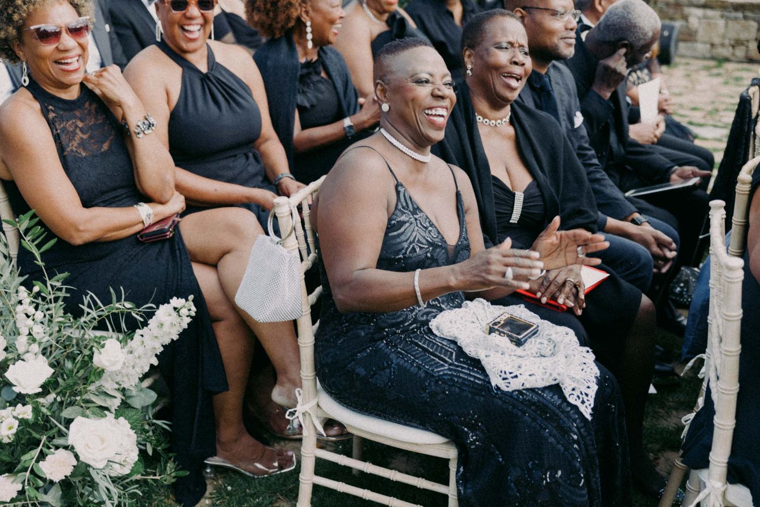 Wedding guests in black attire smile and celebrate during an outdoor ceremony.