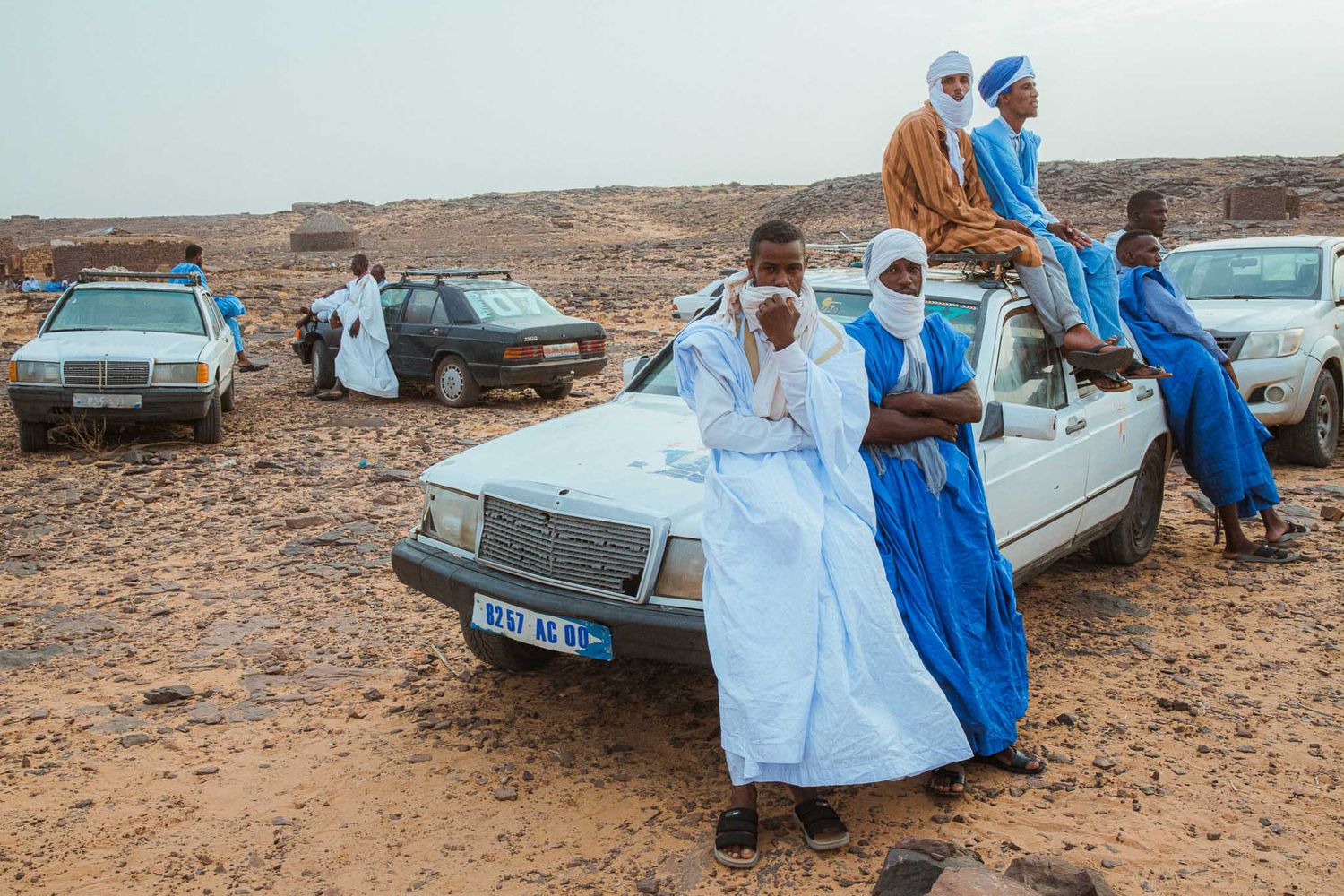 People in traditional blue and white robes sit on the desert ground next to parked vehicles in a remote landscape.