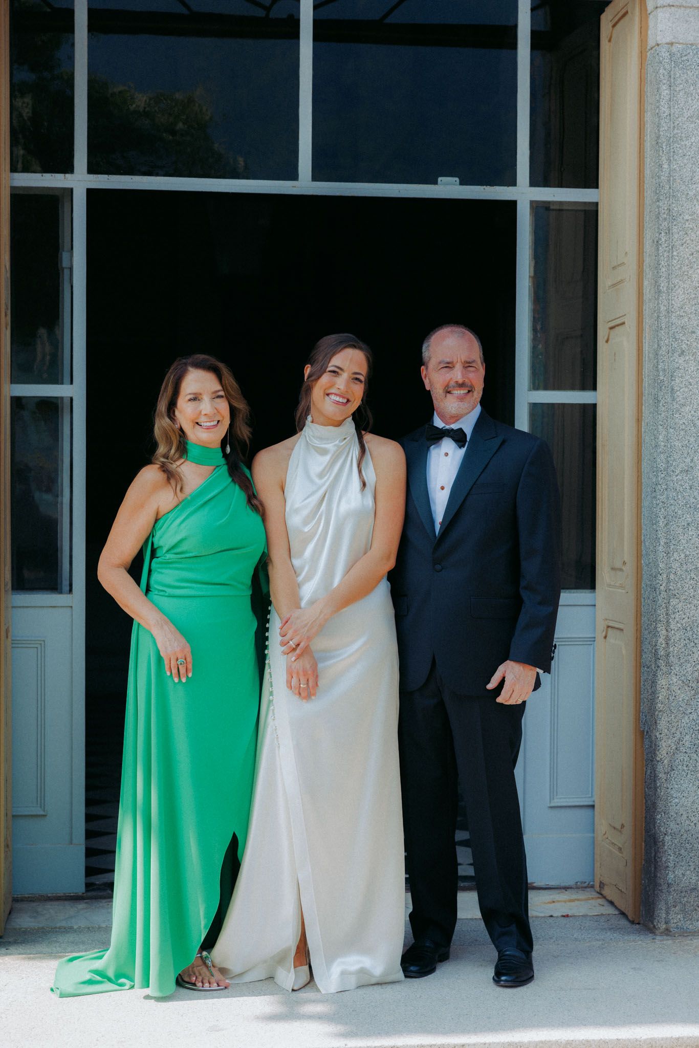 Wedding party in formal attire including green dresses and black tuxedos stand together under an archway.