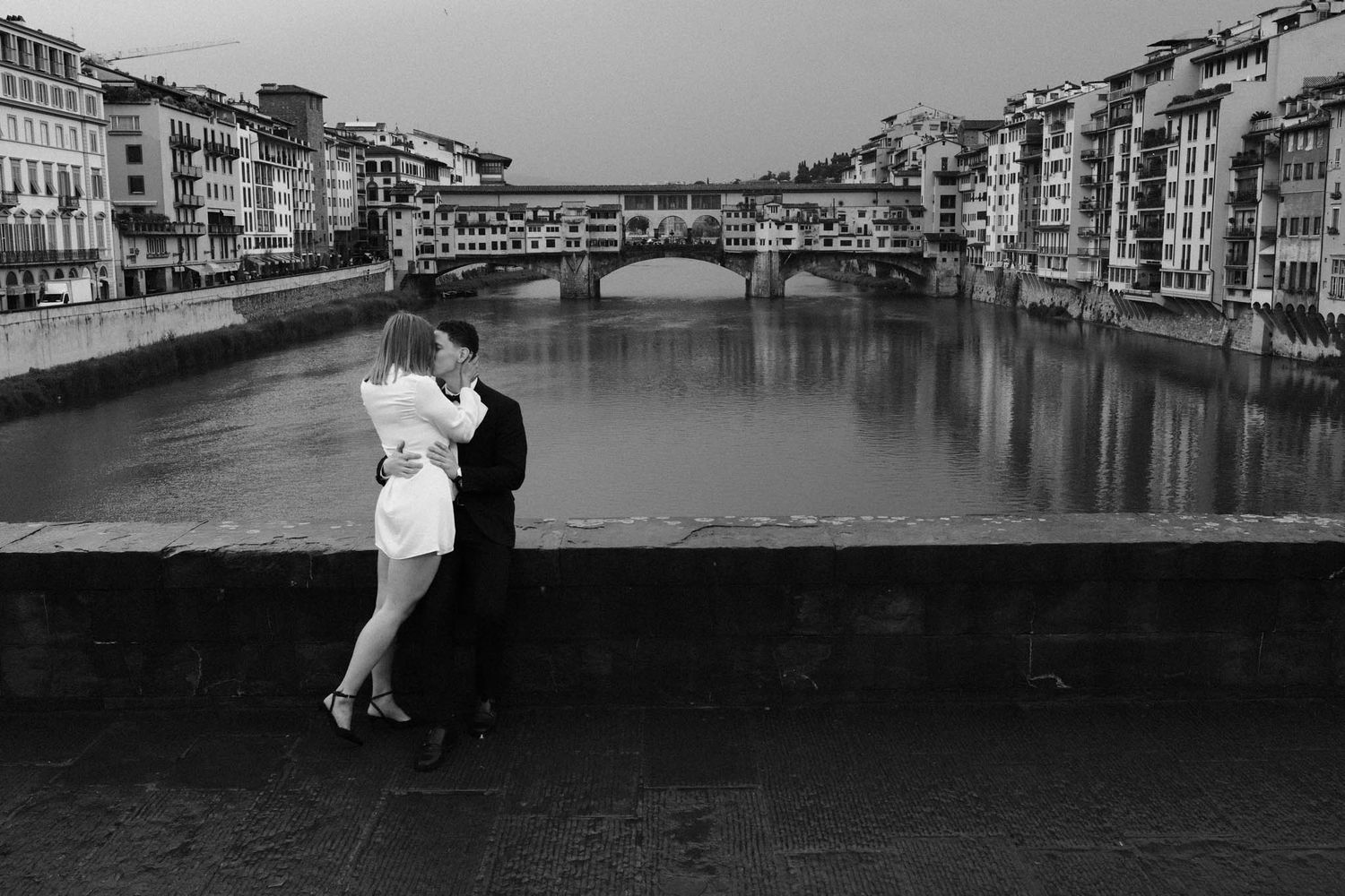 A romantic couple embraces near the iconic Ponte Vecchio bridge in Florence, Italy on a cloudy day.