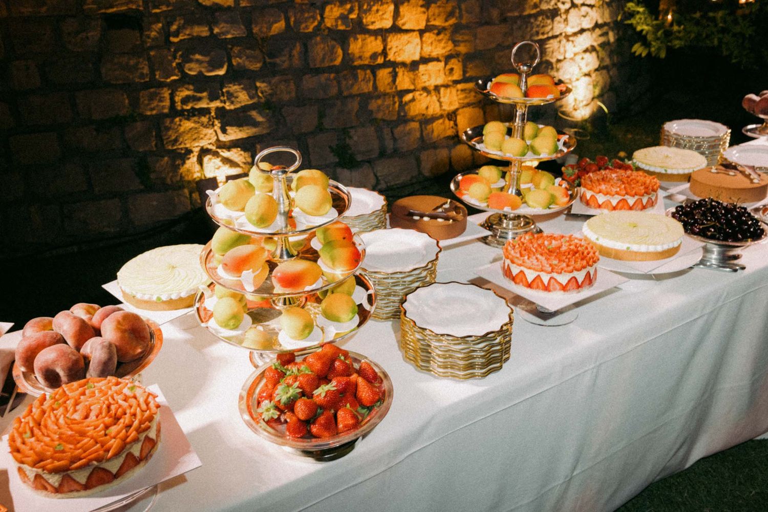An elegant buffet table with tiered dessert stands and platters of pastries illuminated by warm lighting against a brick wall.