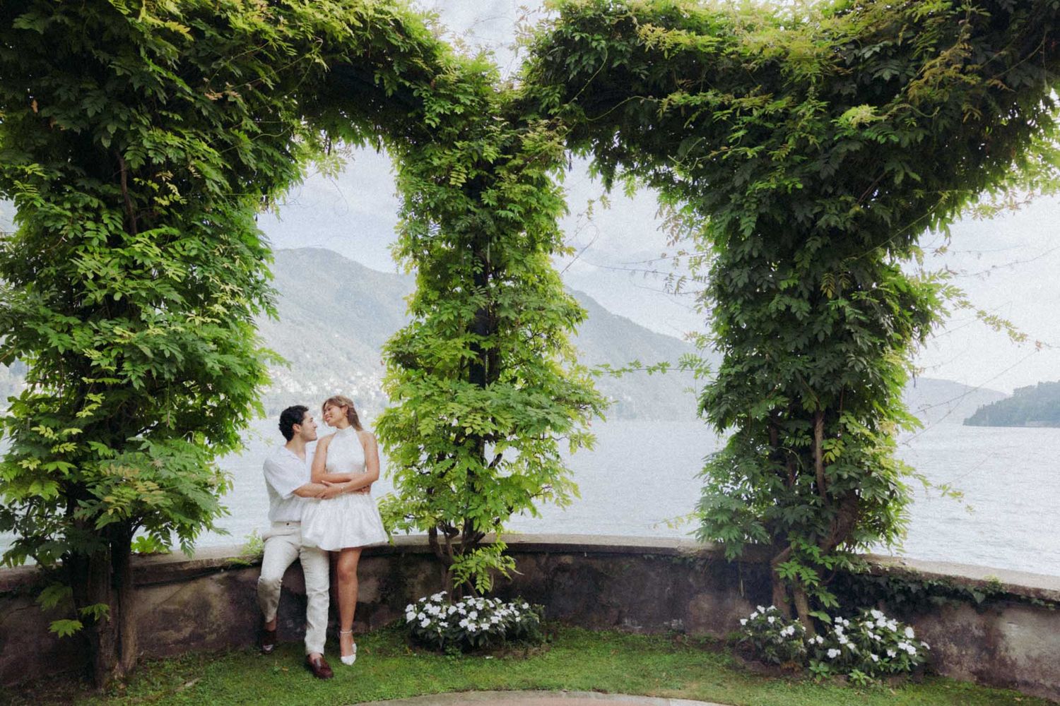 A romantic couple shares an intimate moment in a lush garden setting overlooking a serene lake.