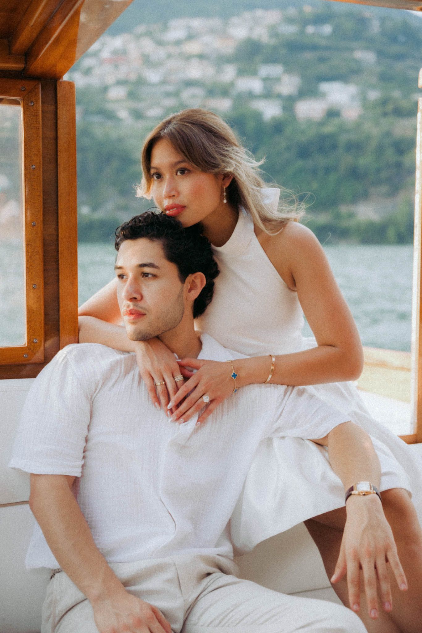 A romantic couple dressed in white poses together on a boat with coastal hills visible in the background.
