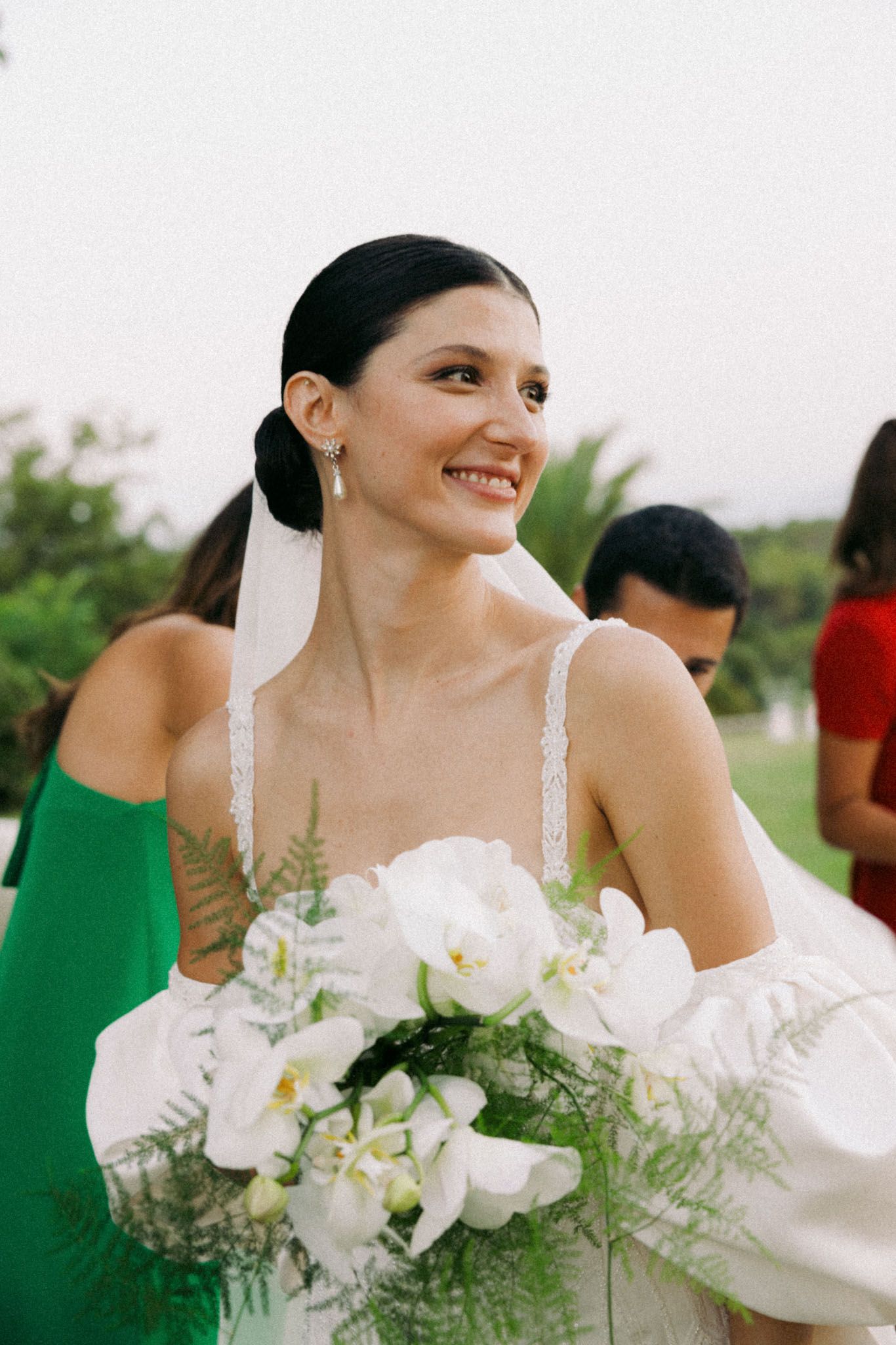 A bride in a white dress holds a bouquet of white flowers and greenery at an outdoor wedding celebration.