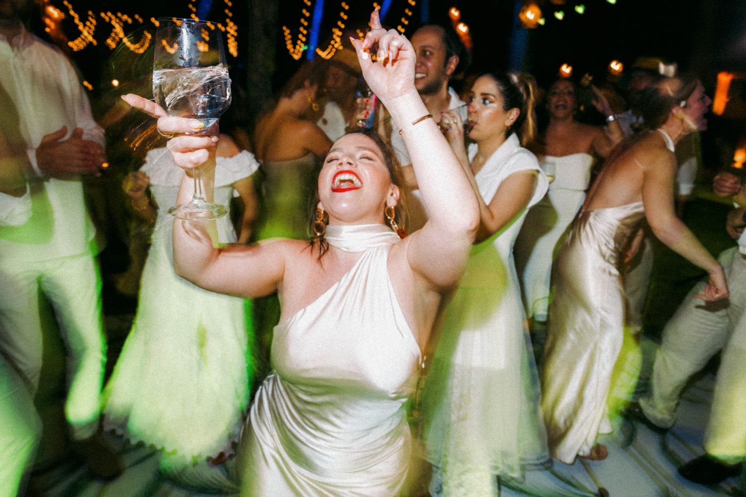 Wedding reception guests in white and mint green dresses dancing joyfully on a crowded dance floor at night.
