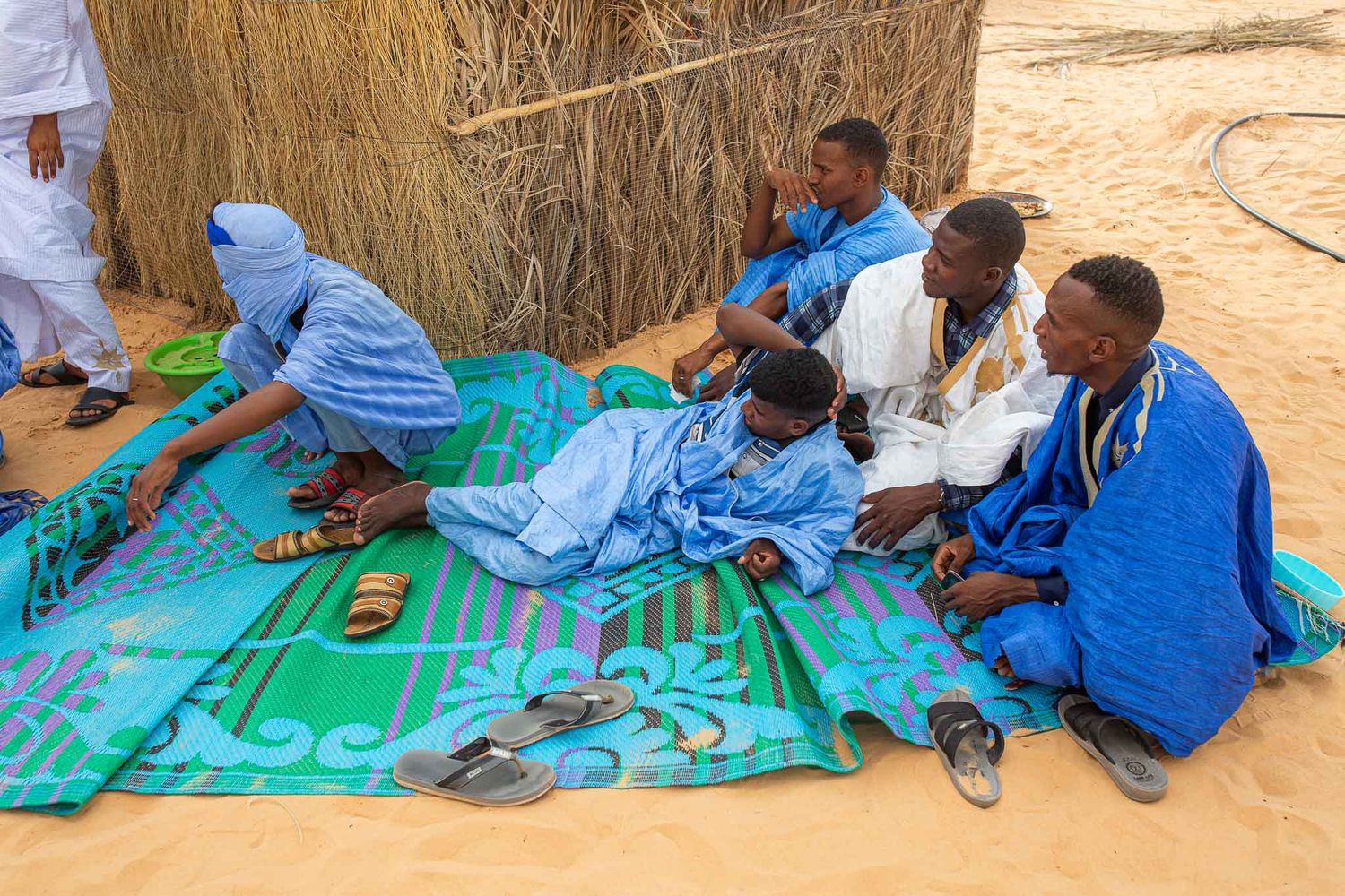 People in traditional blue and white clothing sit on a colorful mat during a gathering in a desert setting.