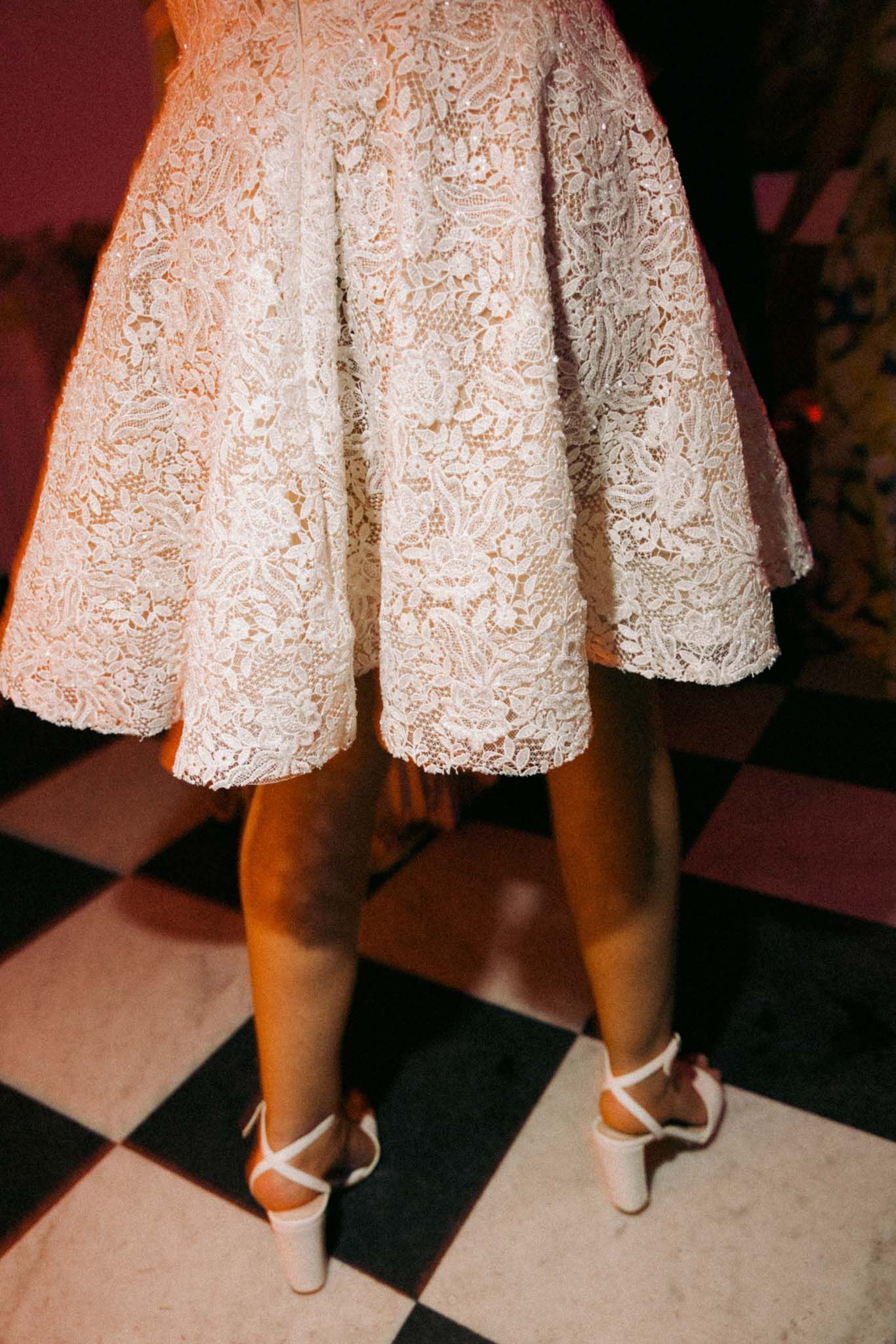 A close-up view of white lace dress hem and heels on a black and white checkered floor.