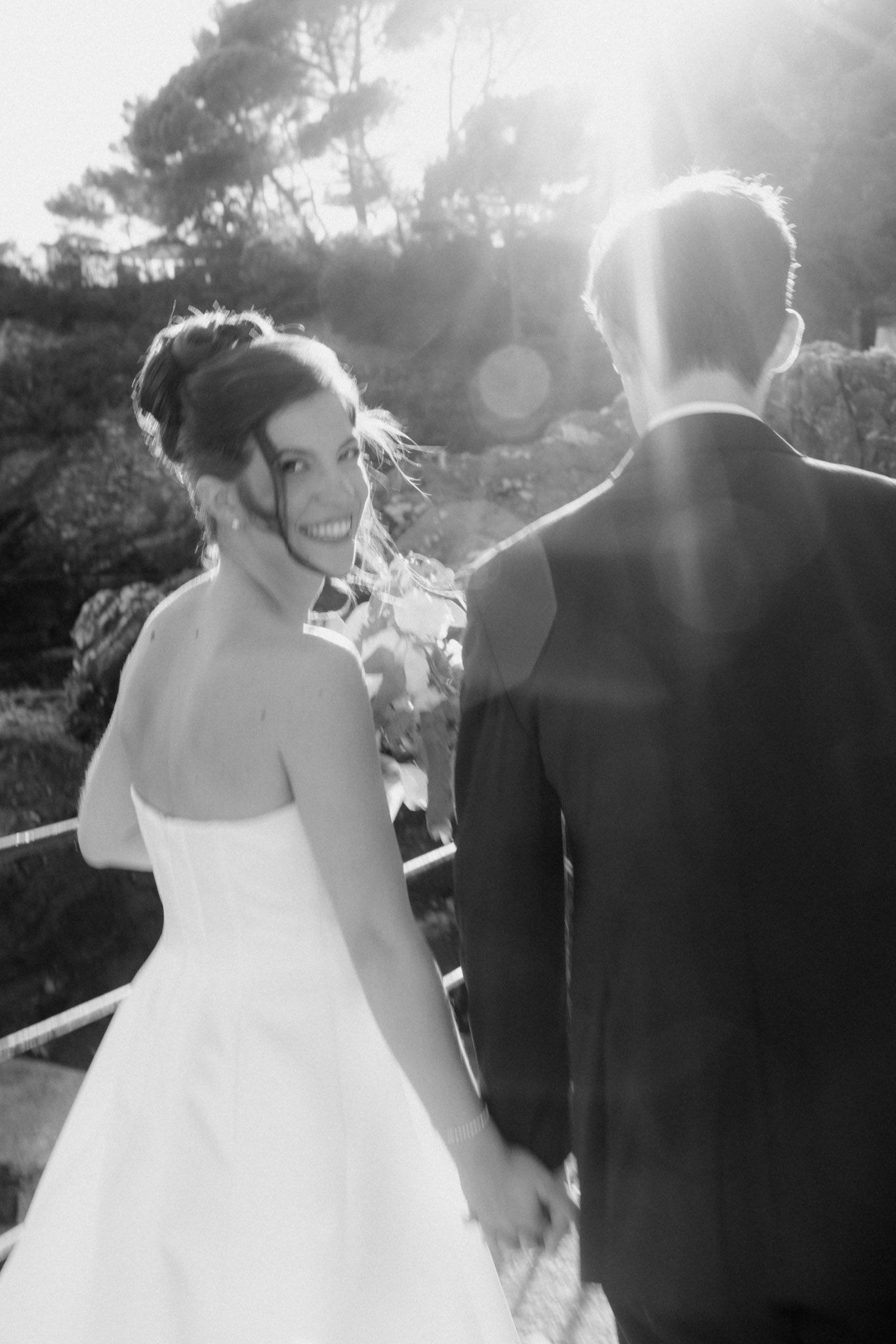 A bride and groom walk together outdoors in black and white wedding photos with sunlight streaming through trees.