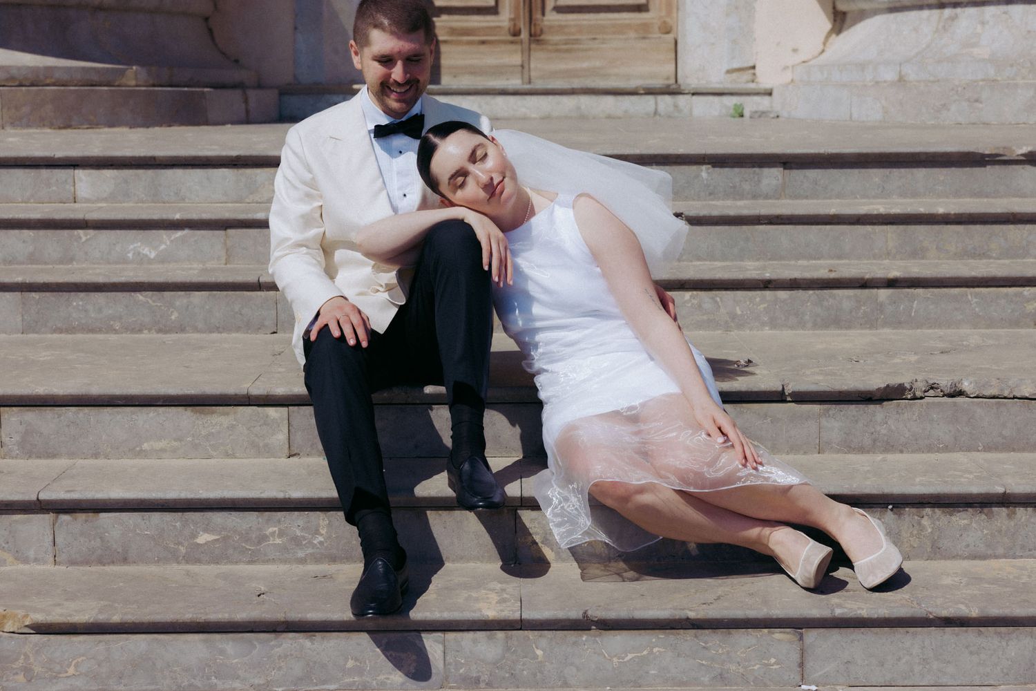A romantic wedding couple shares tender moments while sitting on stone steps in an elegant outdoor setting.