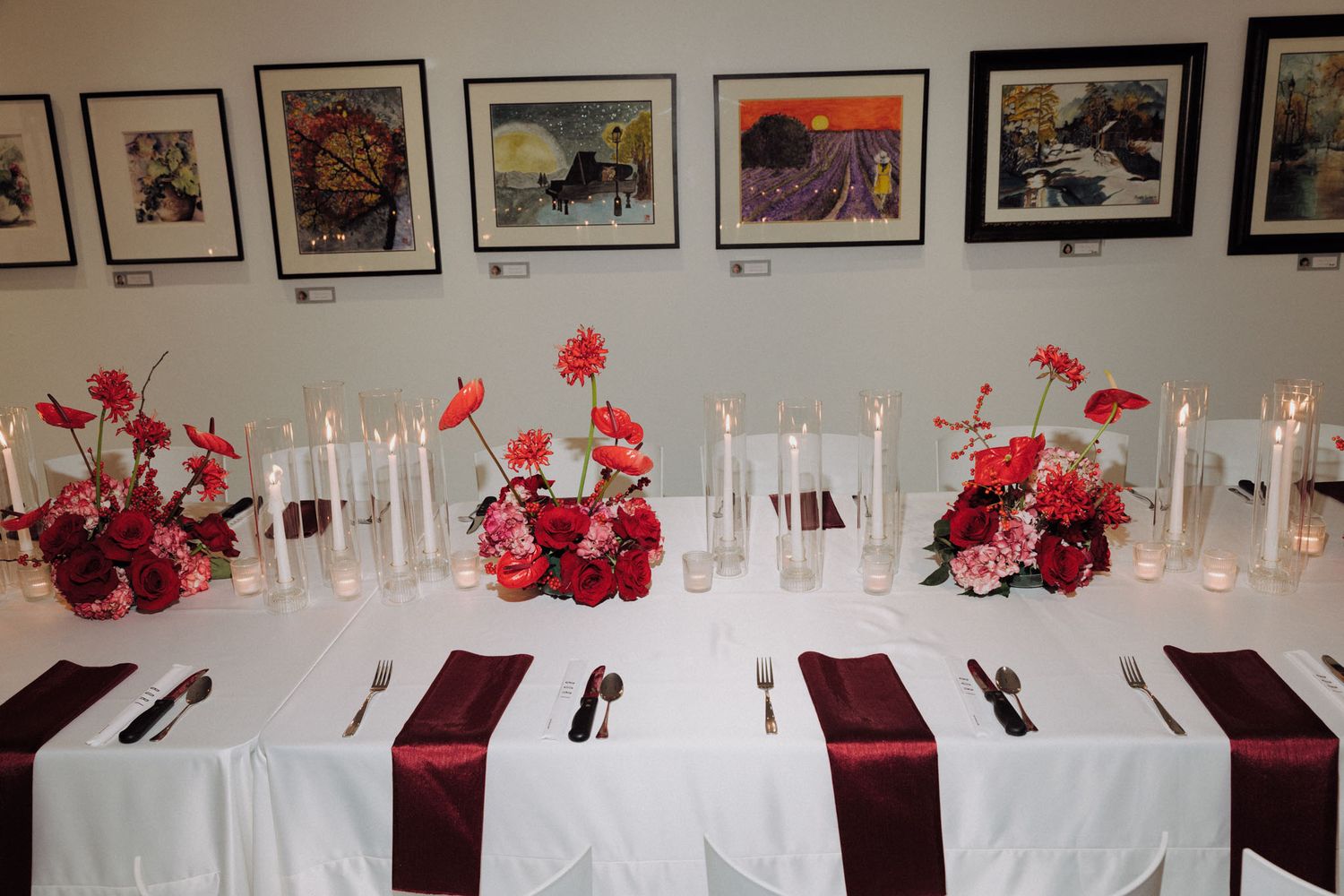 Elegant wedding reception table decorated with red roses and white candles in a gallery-style venue with artwork on walls.
