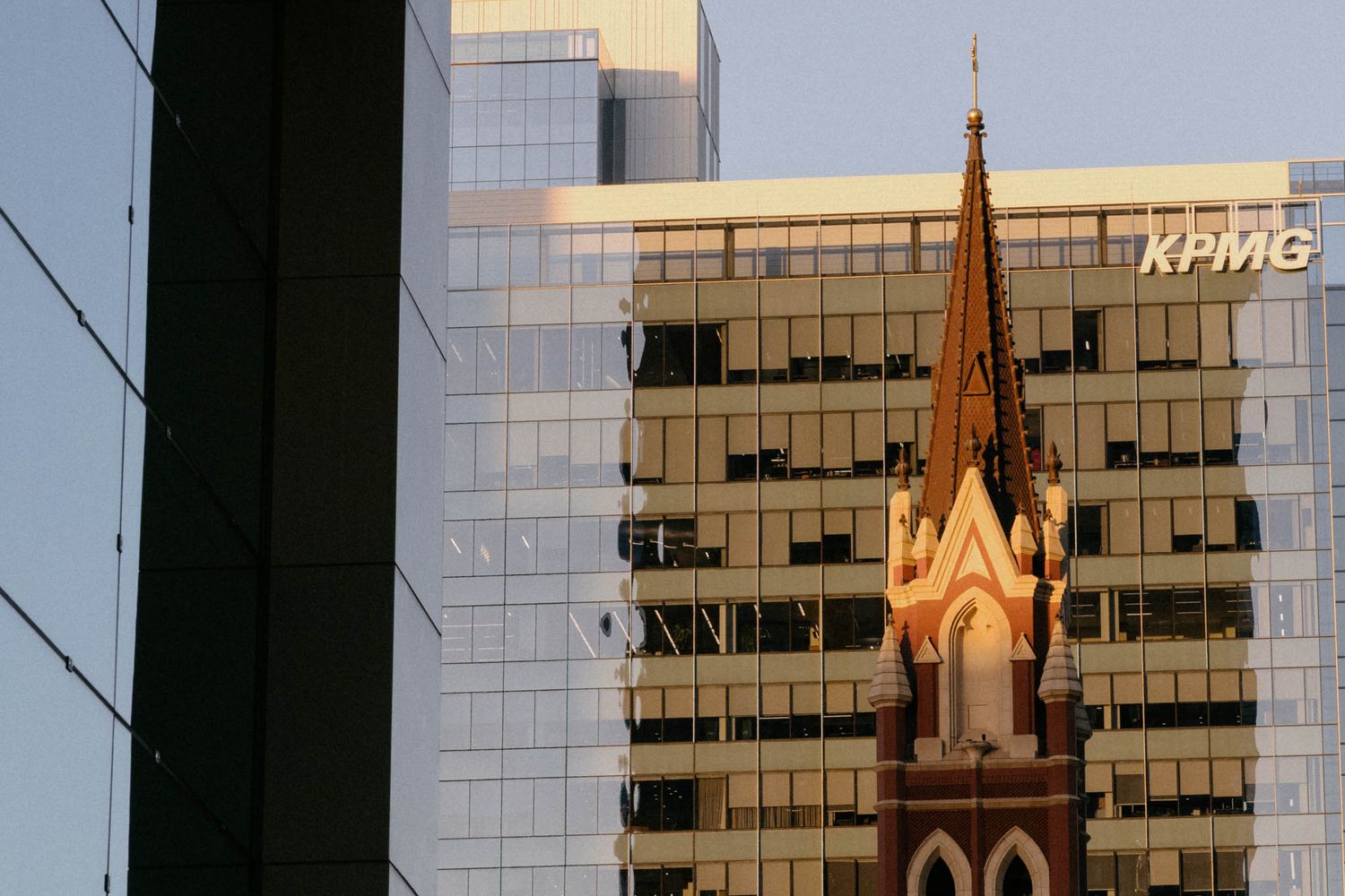 Modern glass office buildings reflect sunset light alongside historic church spires in an urban cityscape.