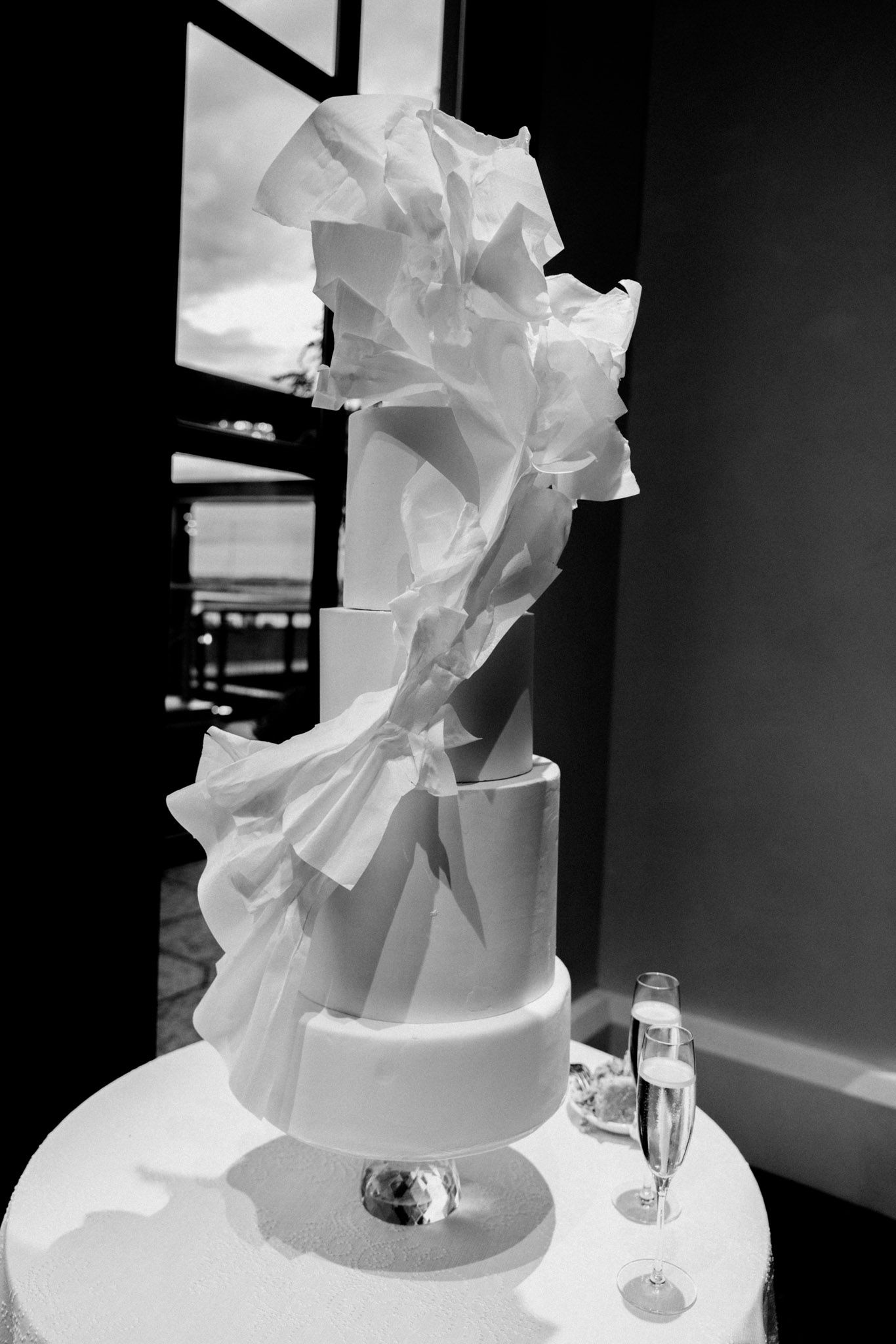 A dramatic black and white photo of a modern white wedding cake with flowing ruffled details and elegant tiered design.