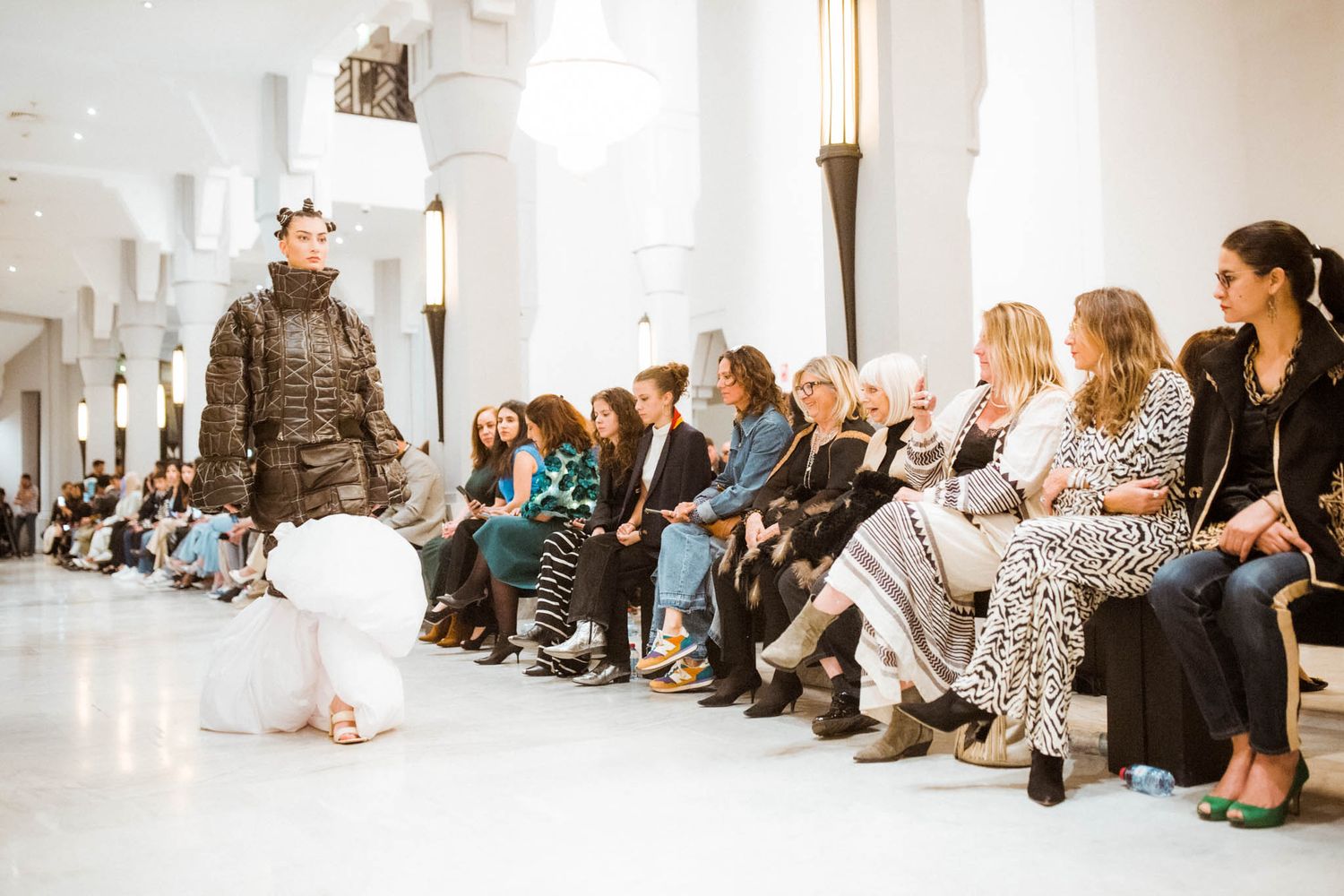 People seated in rows along a white wall during an indoor event with bright lighting and minimalist decor.