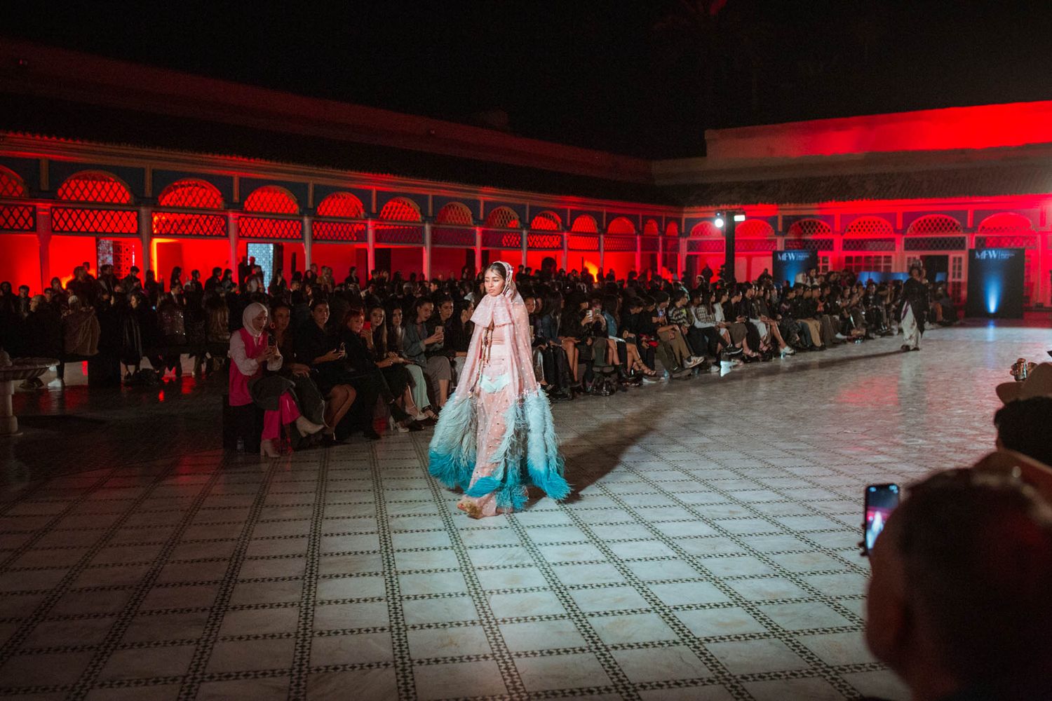 Performance venue illuminated in dramatic red lighting with audience seating visible in the background.