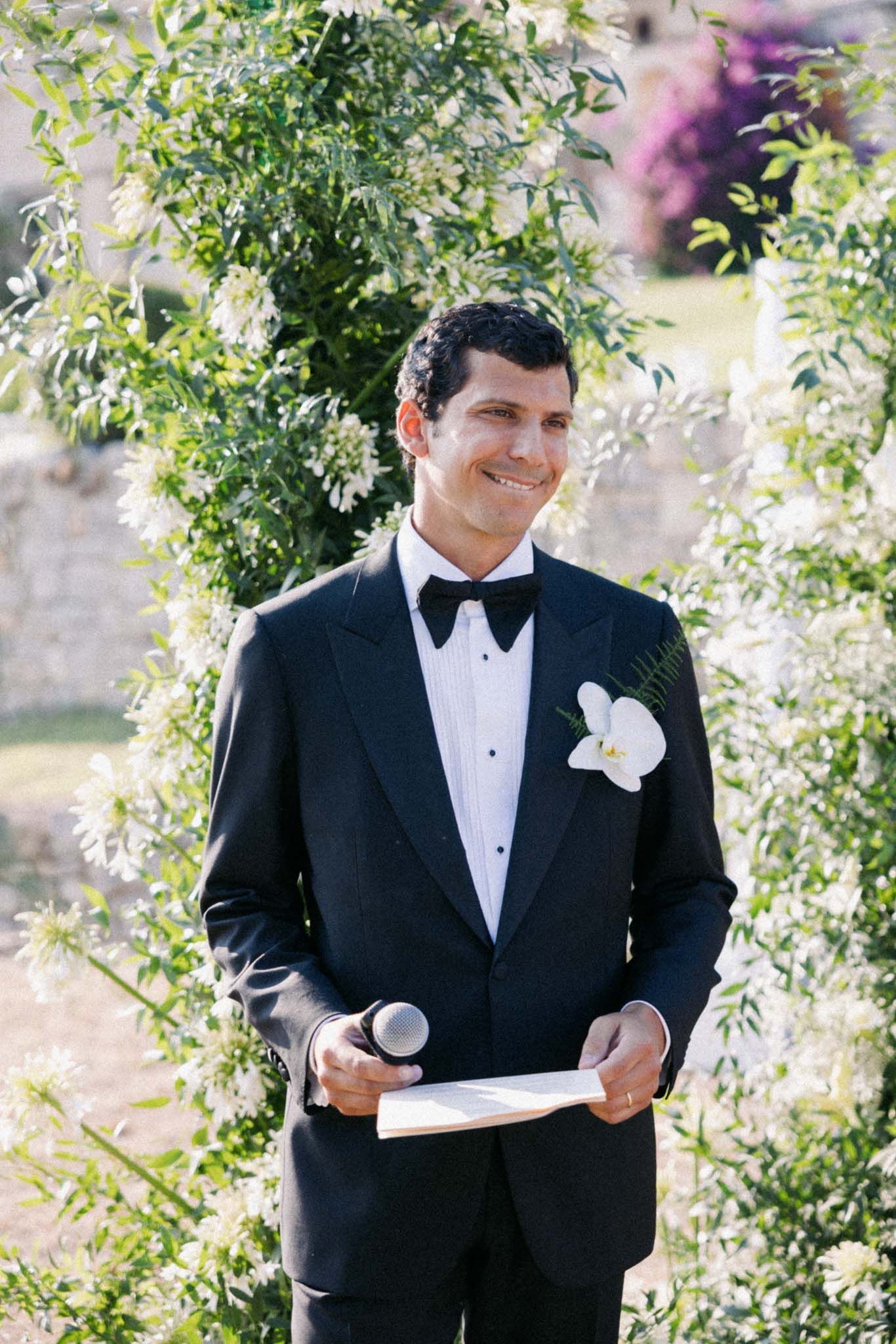 A person in a black tuxedo with a white boutonniere smiles while holding papers at an outdoor wedding ceremony.