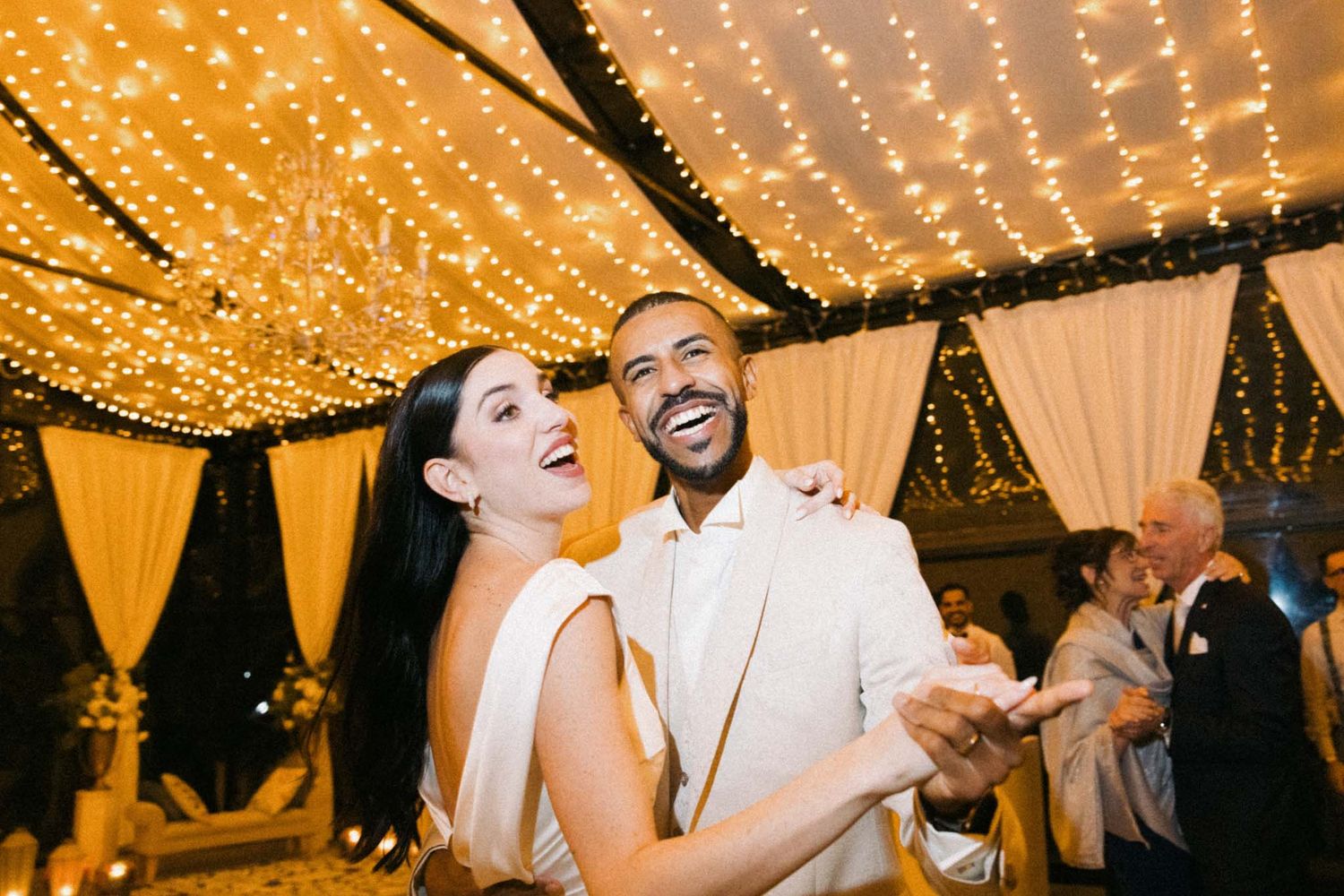 A couple dances joyfully under a canopy of warm glowing string lights at an elegant wedding reception.
