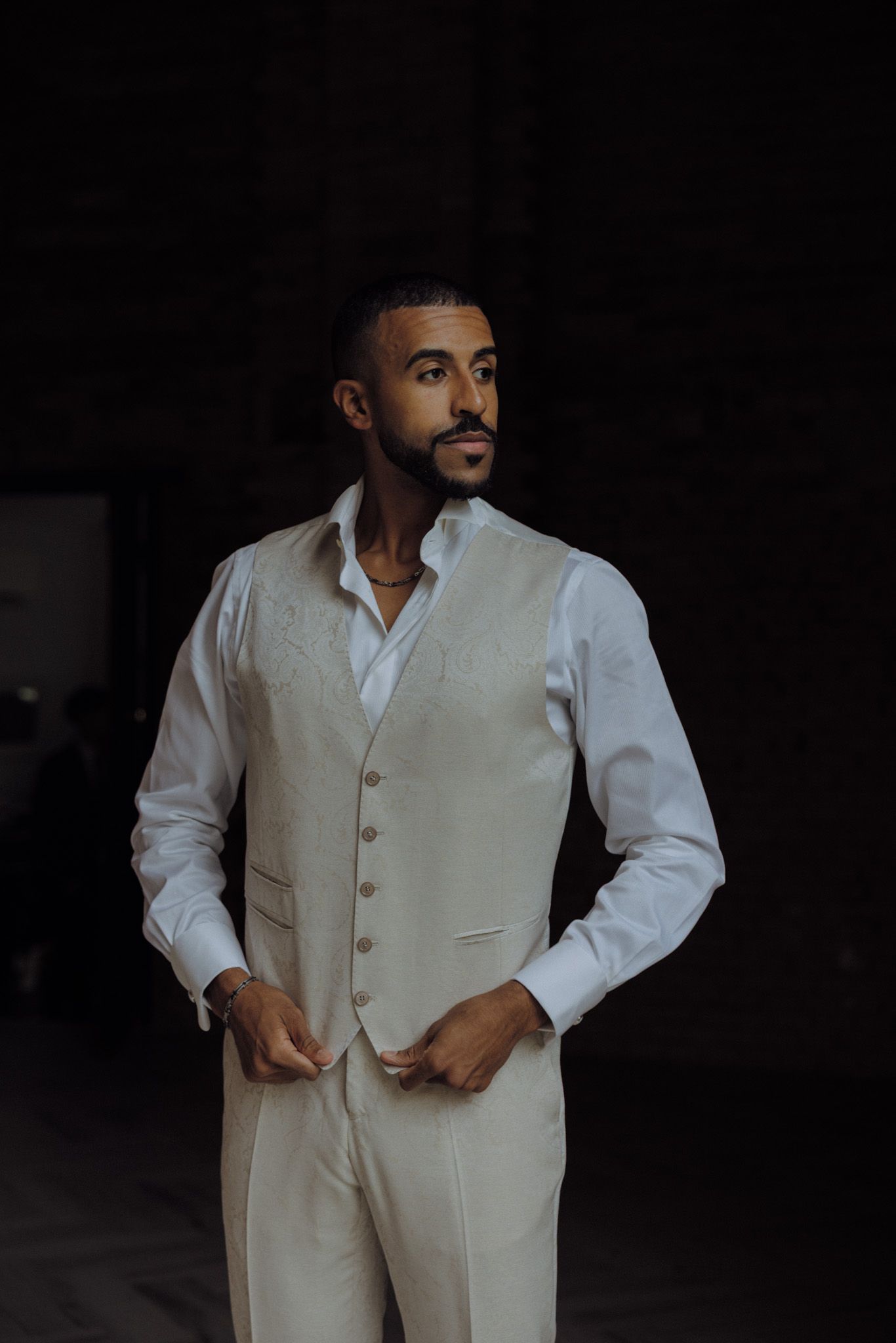 A well-dressed person in a light-colored vest and white shirt stands confidently against a dark background.