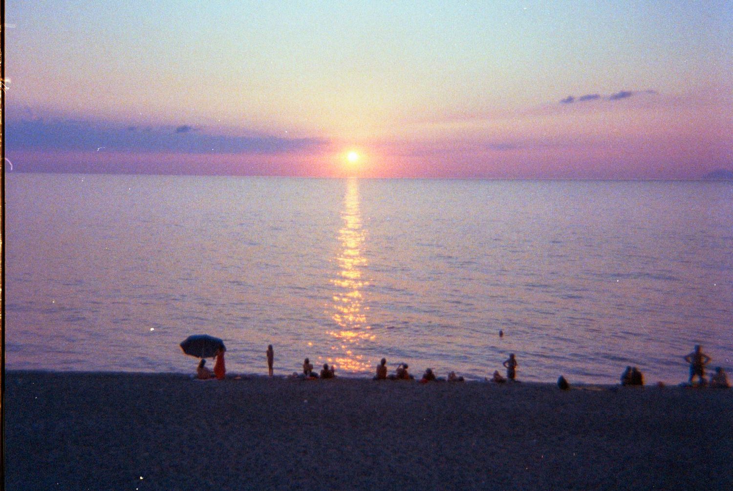 Silhouetted figures gather along a beach at sunset with vibrant orange and purple sky reflecting on calm ocean waters.