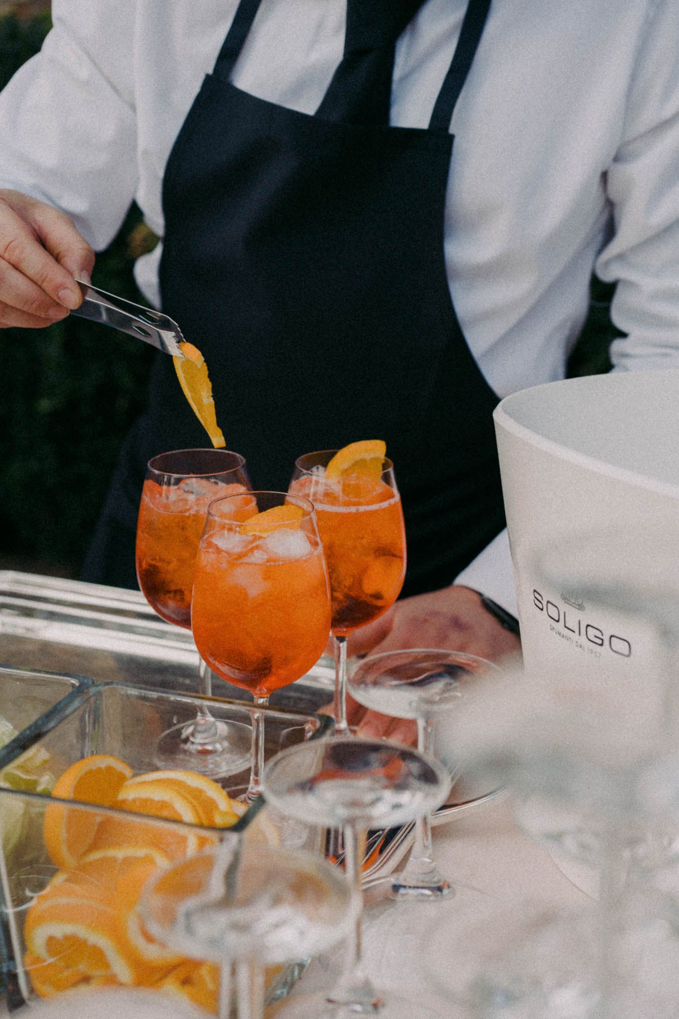 A bartender garnishes an Aperol Spritz cocktail with fresh orange slices on a white marble bar counter.