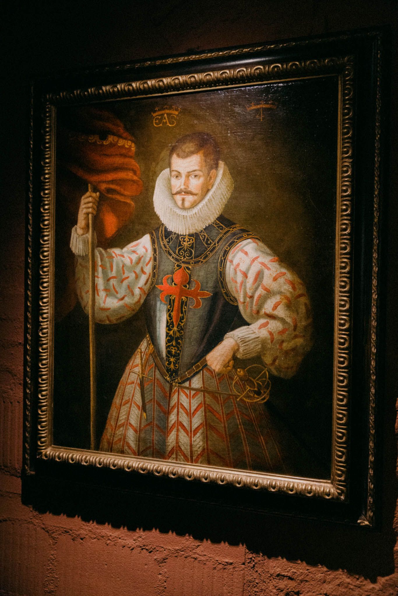 Four dark classical portrait paintings in ornate gold frames hang on a textured wall in dim lighting.