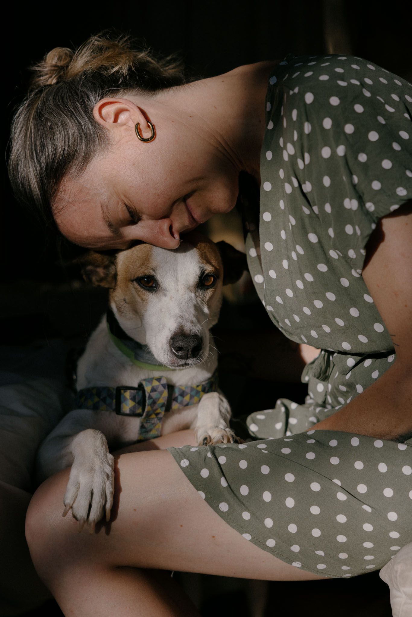 A small white dog receives affectionate attention while resting on a dark surface in a dimly lit setting.