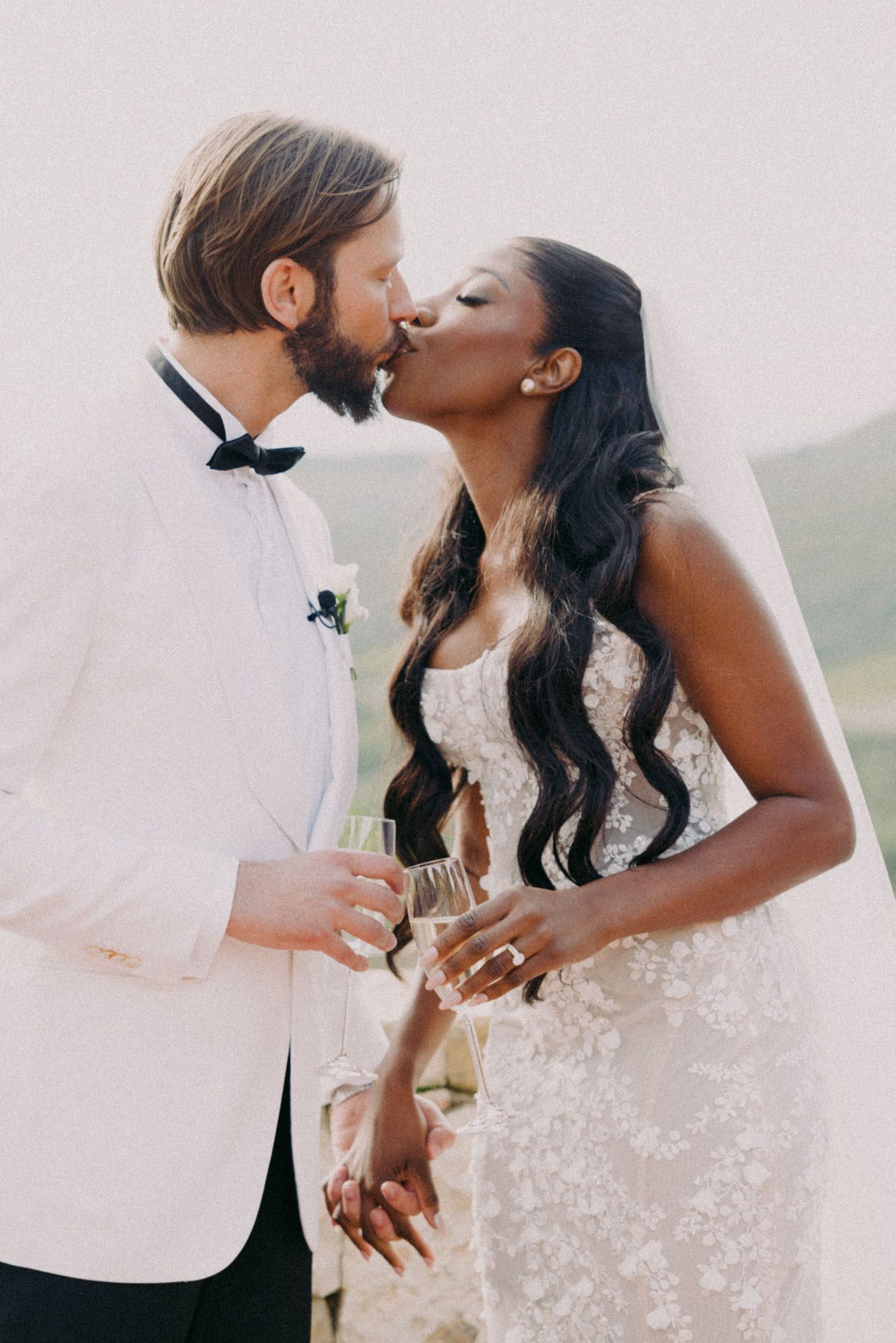 A romantic wedding couple sharing a kiss outdoors, with the bride in a lace dress and groom in a white dinner jacket.