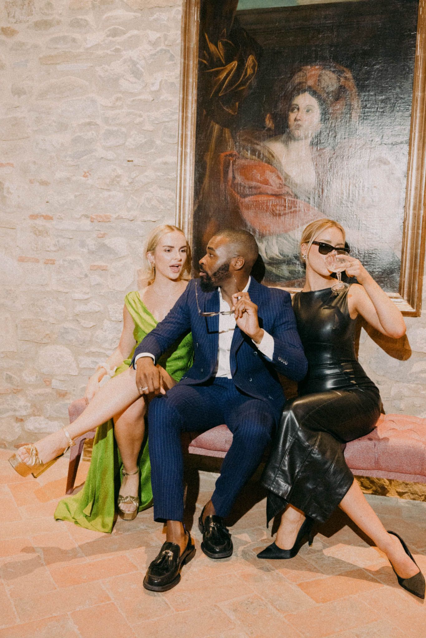 People in formal attire sit together on a tiled floor against a brick wall with framed artwork.