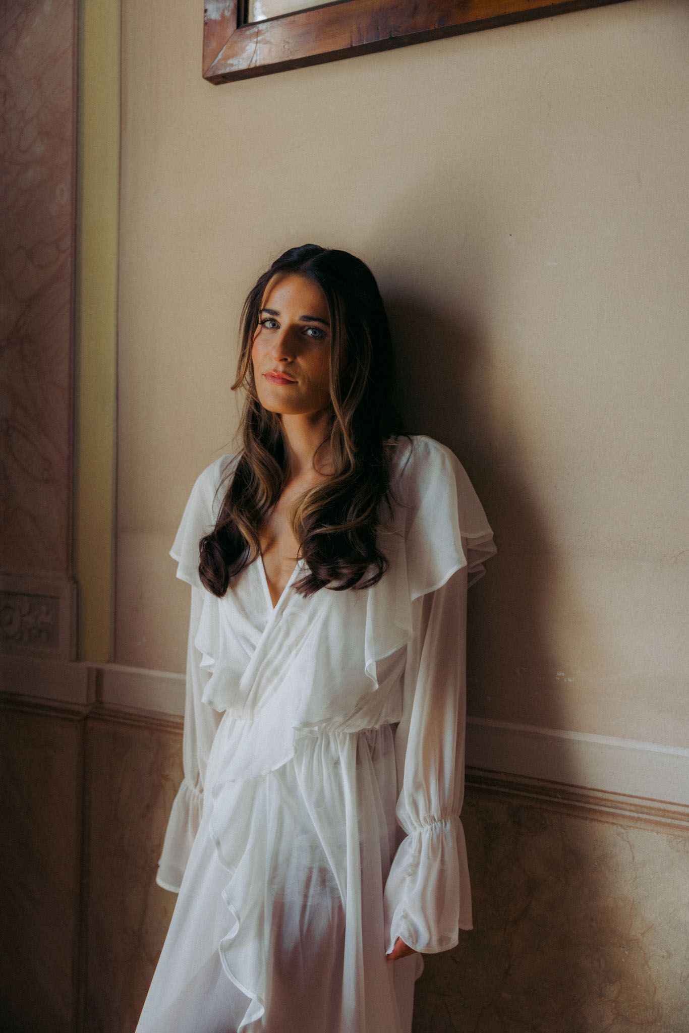 A figure in a flowing white robe with ruffled sleeves stands against a beige wall in moody lighting.