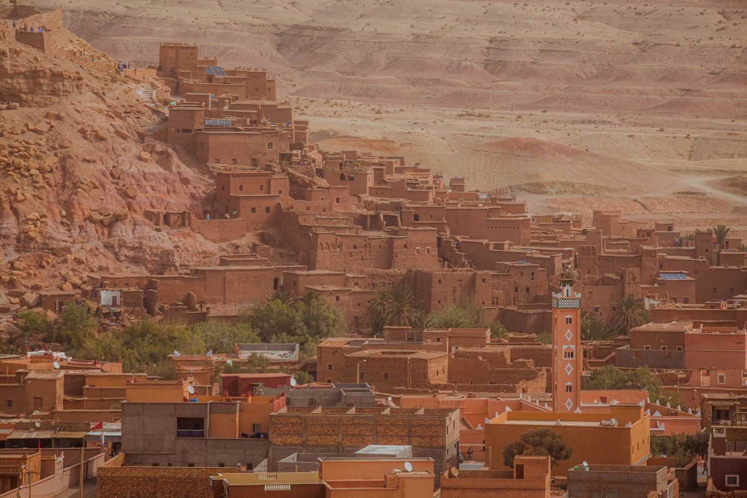 Ancient clay buildings and structures line the hillside in a traditional Moroccan village under a cloudy sky.