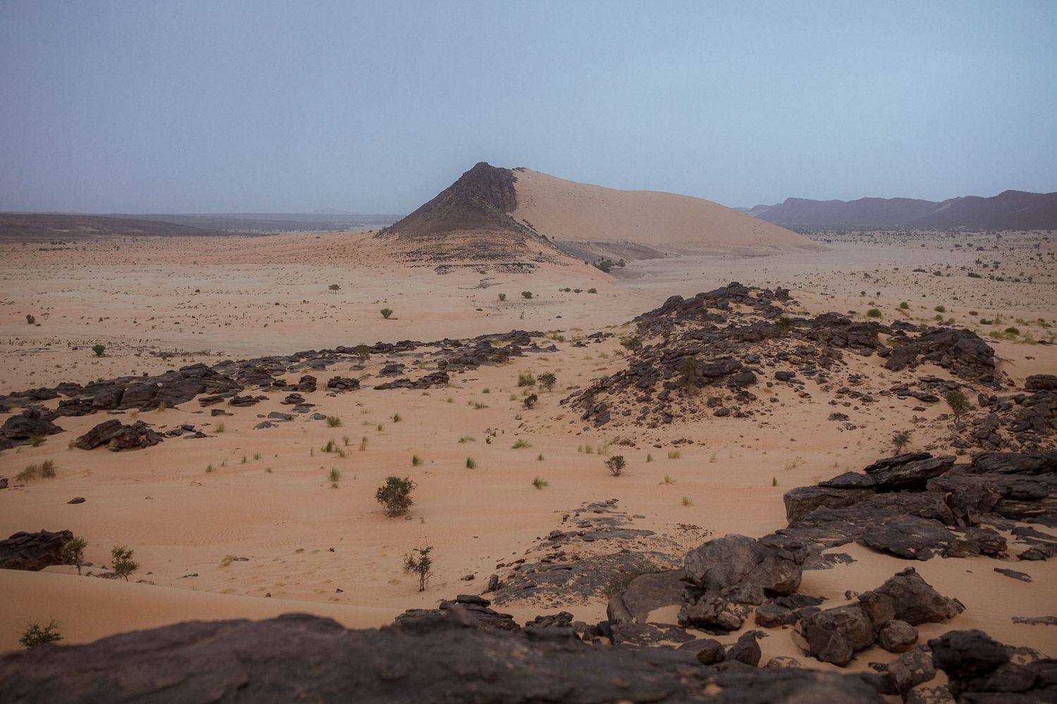 Desert landscape with sand dunes and rocky outcrops under cloudy sky.