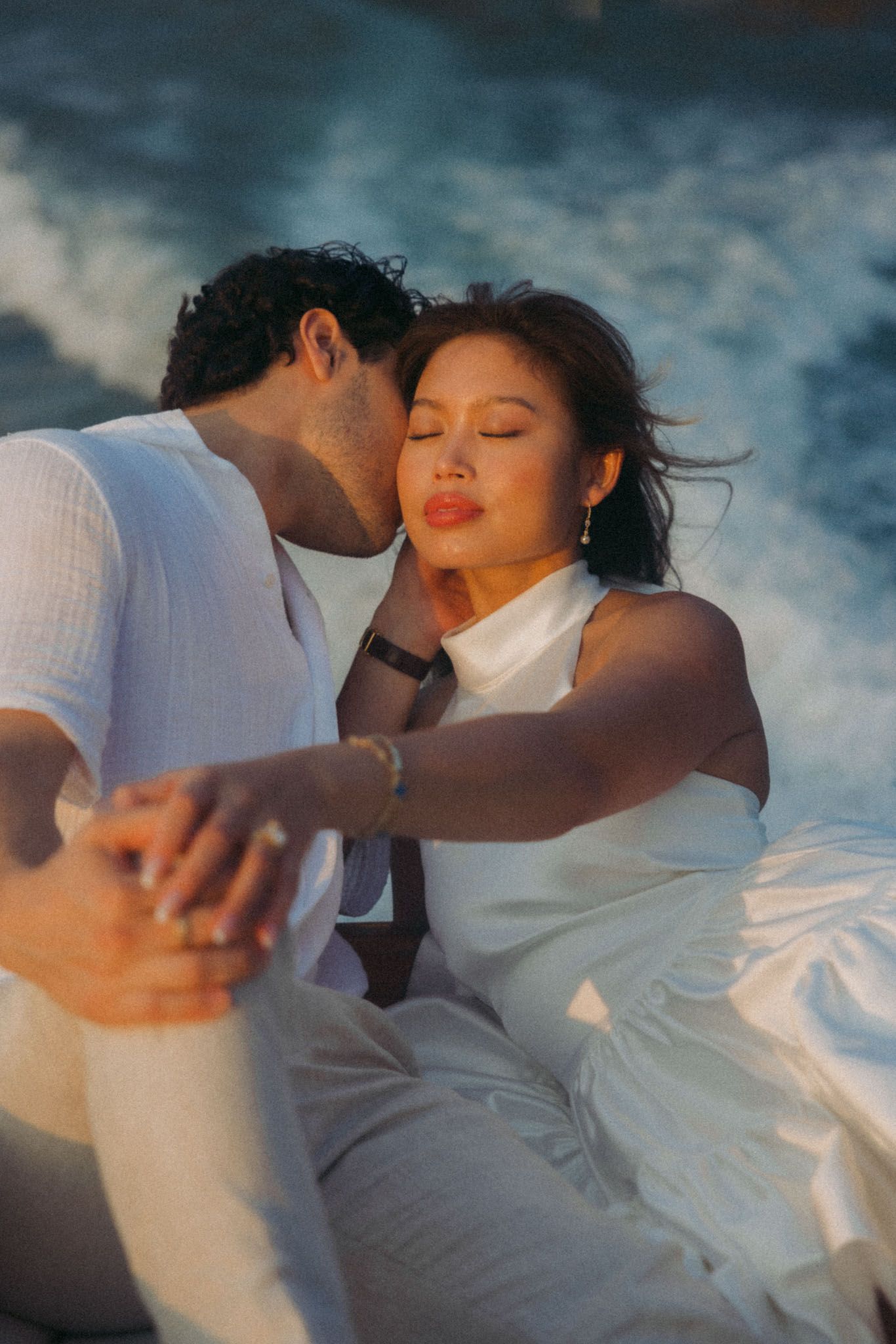 A romantic couple in white clothing embraces passionately against a moody blue sky background.