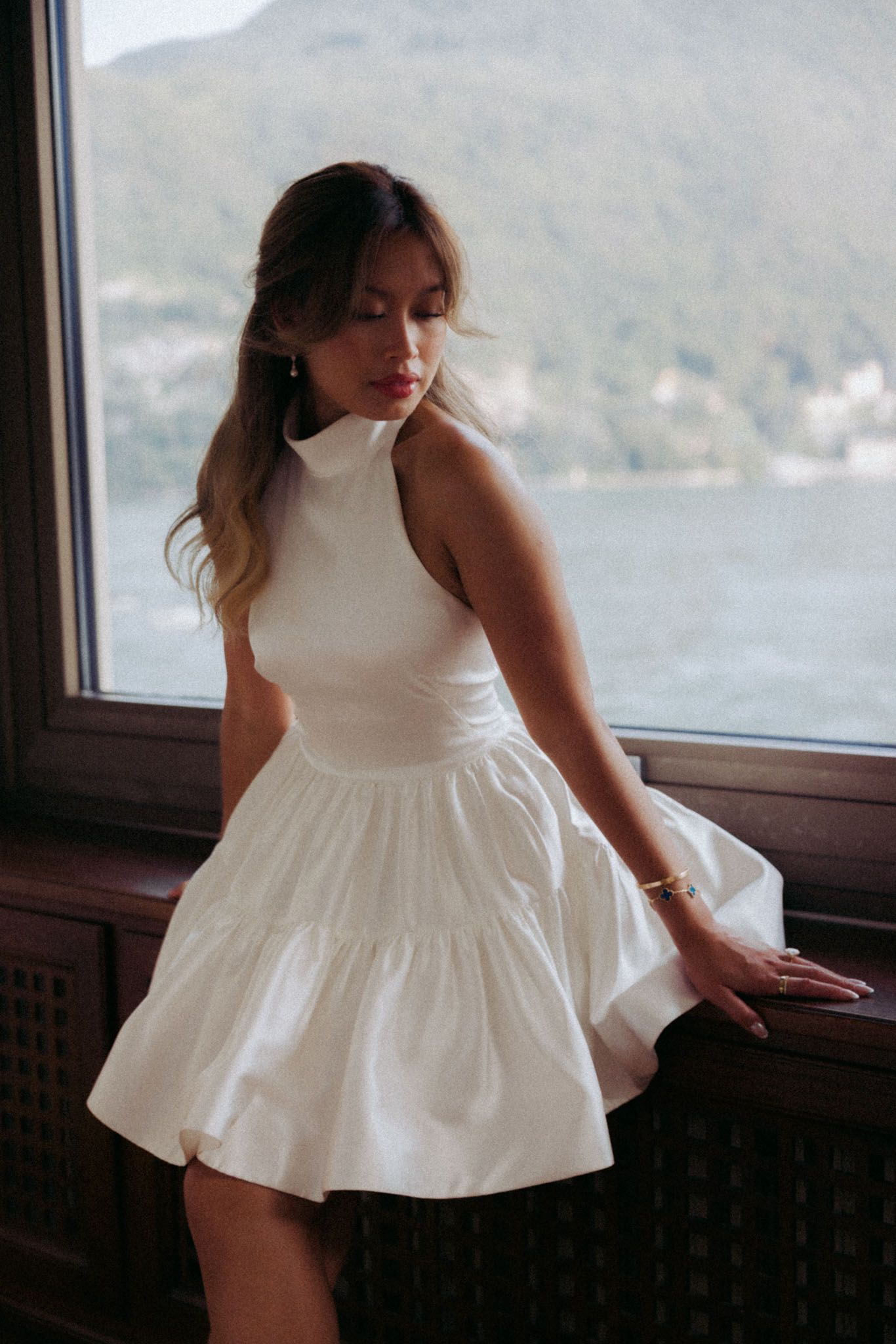 A figure in a flowing white dress poses by a window with a scenic mountain and water view in the background.