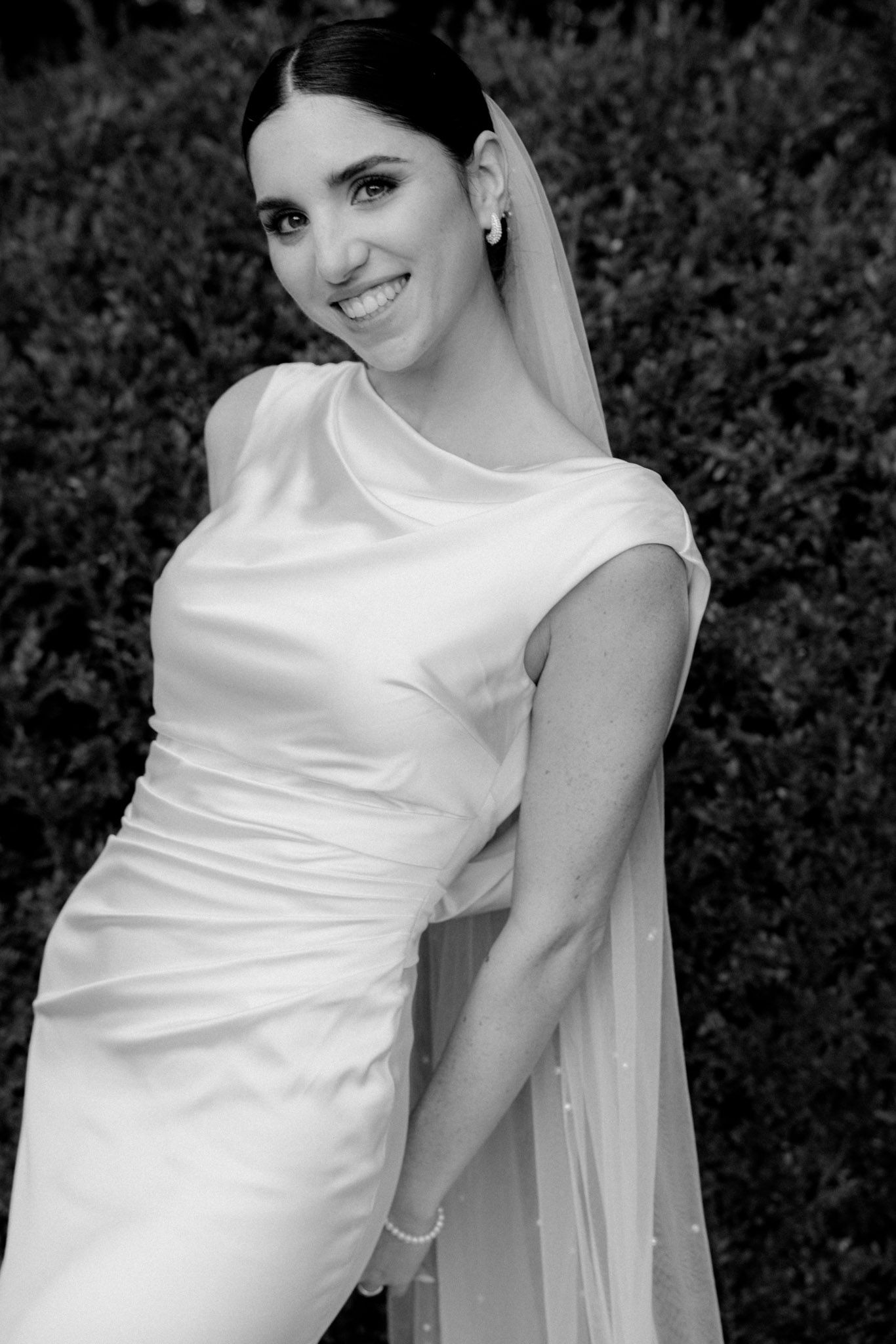 Black and white portrait of a bride in an elegant fitted satin wedding dress with cap sleeves and flowing veil.