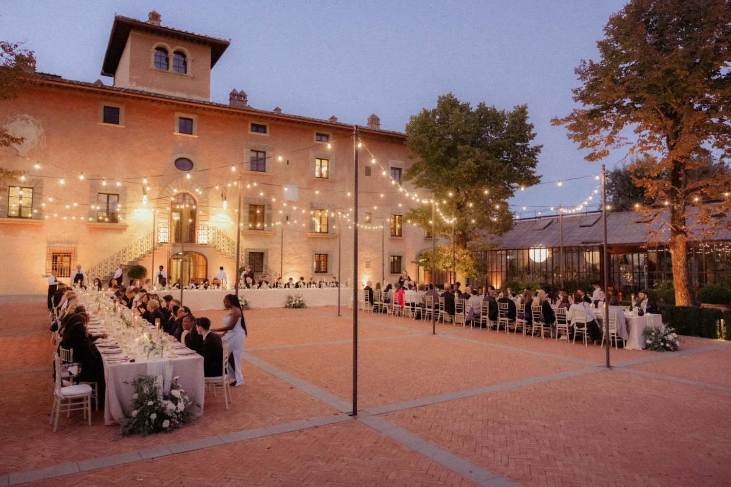 Panoramic view of a courtyard with string lights and outdoor seating at dusk creating a warm, inviting atmosphere.