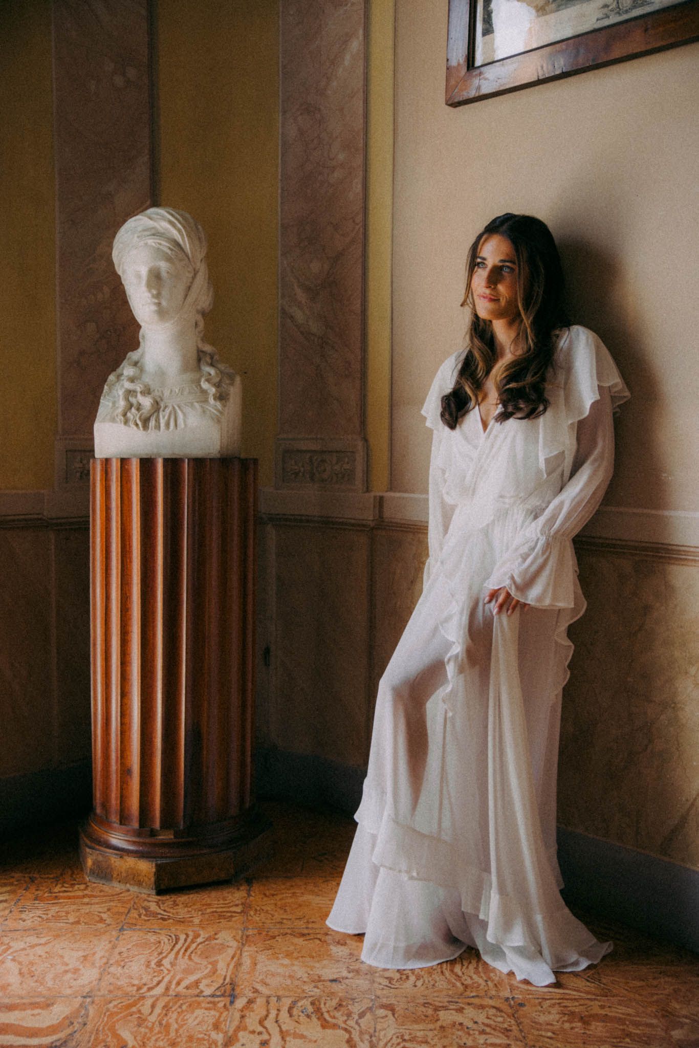 Moody indoor photoshoot featuring a figure in white clothing posed alongside classical busts and wooden panels.