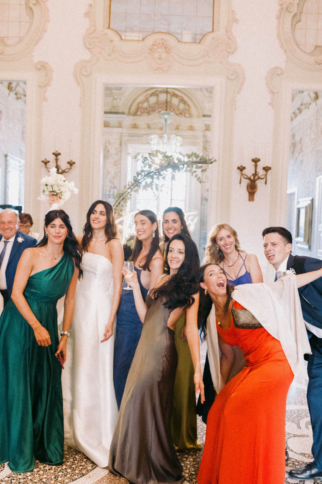 A group of wedding guests in colorful formal dresses laugh and pose together in an elegant ballroom setting.