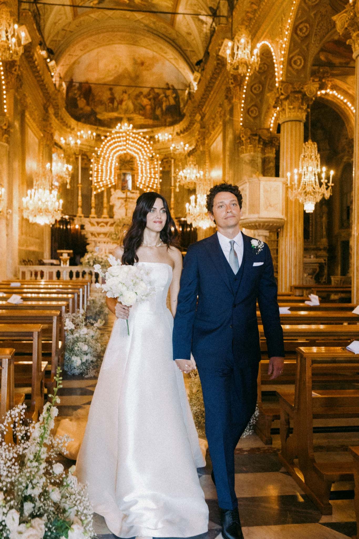 A couple poses in an elegant church interior with ornate chandeliers and baroque decor during a wedding ceremony.