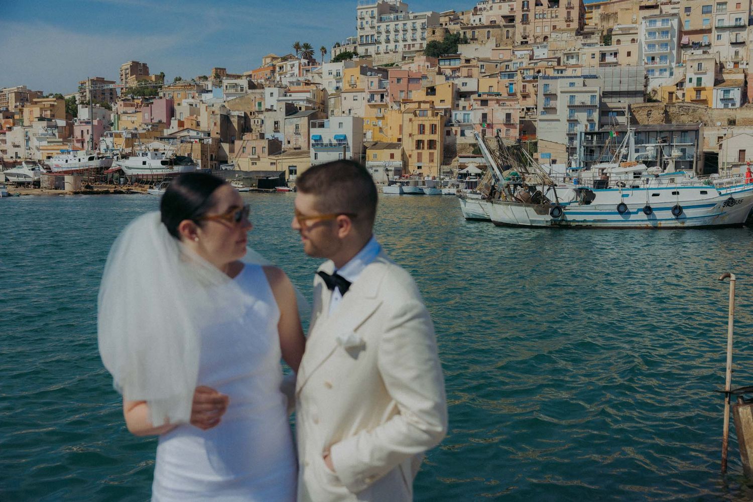 A couple in wedding attire shares a romantic moment on a boat with colorful coastal buildings in the background.