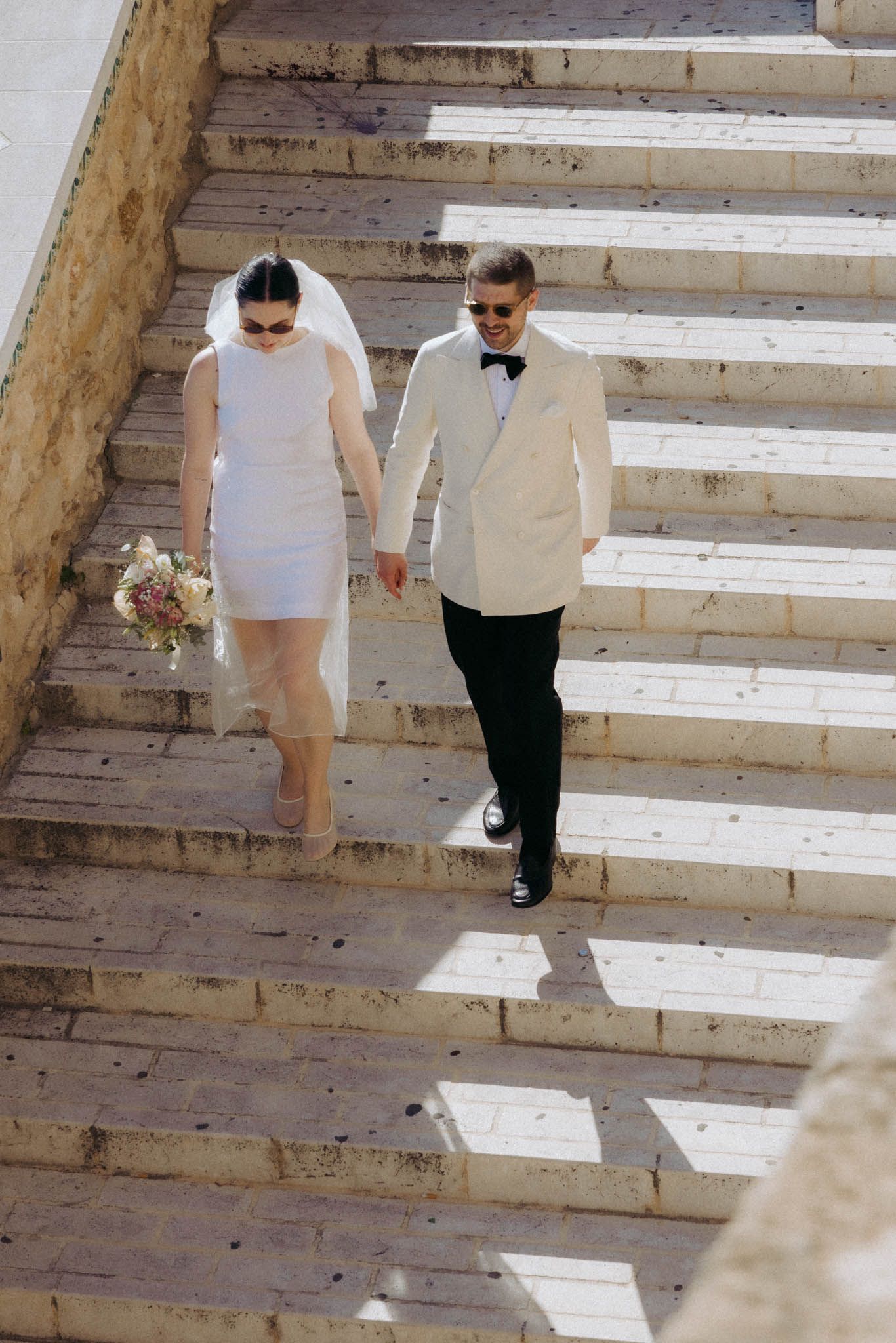Couple walking down sunlit stone steps in formal attire during a wedding photoshoot.