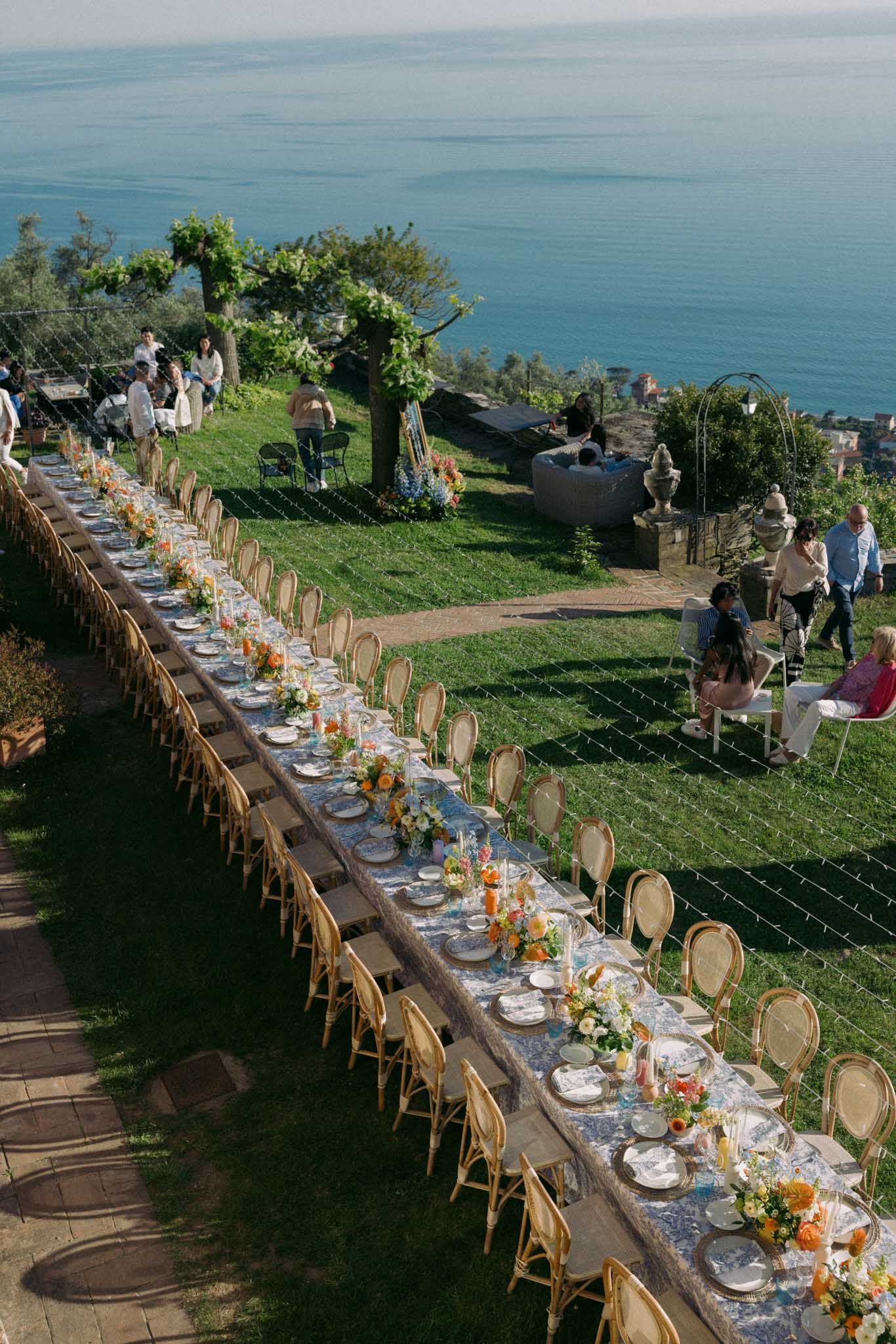 Long elegant dining table set up on a cliff overlooking the ocean with floral centerpieces and wooden chairs.