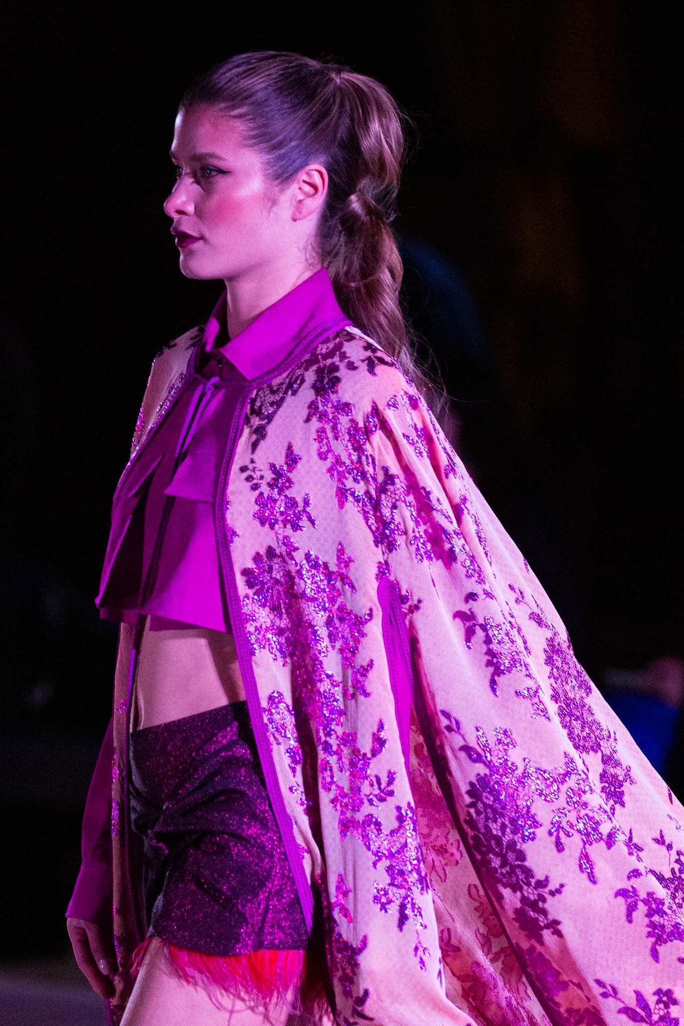 Model walks runway in floral print pink robe with dramatic lighting at fashion show.