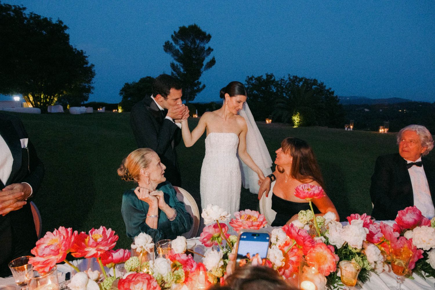 People gather at an outdoor dinner party with vibrant coral and pink floral centerpieces on long tables at dusk.