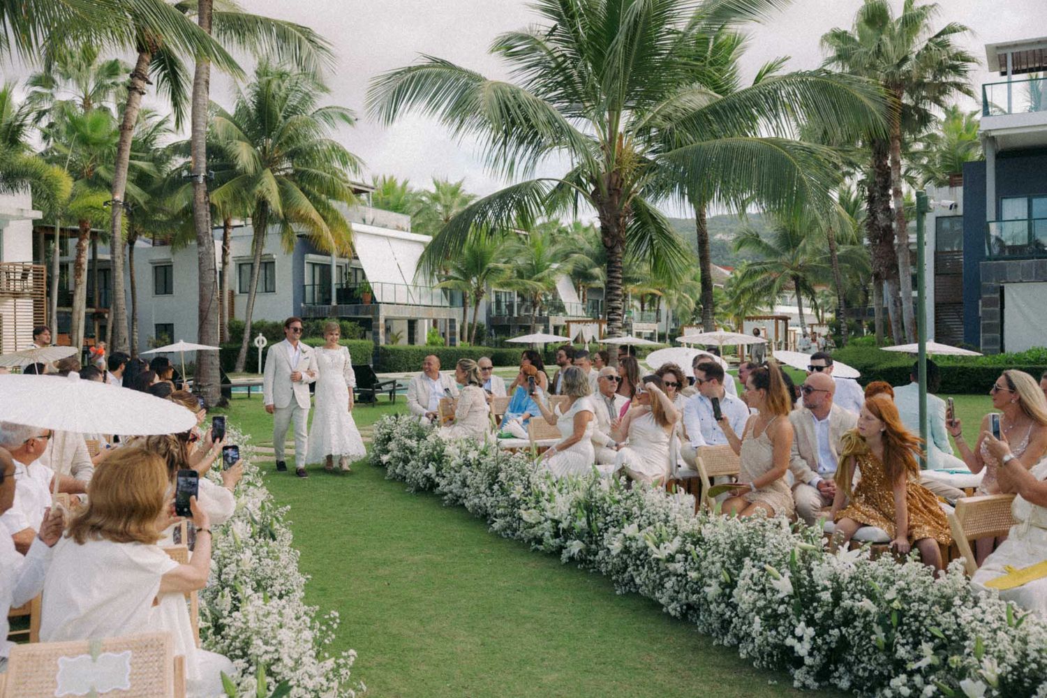 Outdoor wedding ceremony setup with white floral arrangements along the aisle and palm trees in a tropical garden setting.
