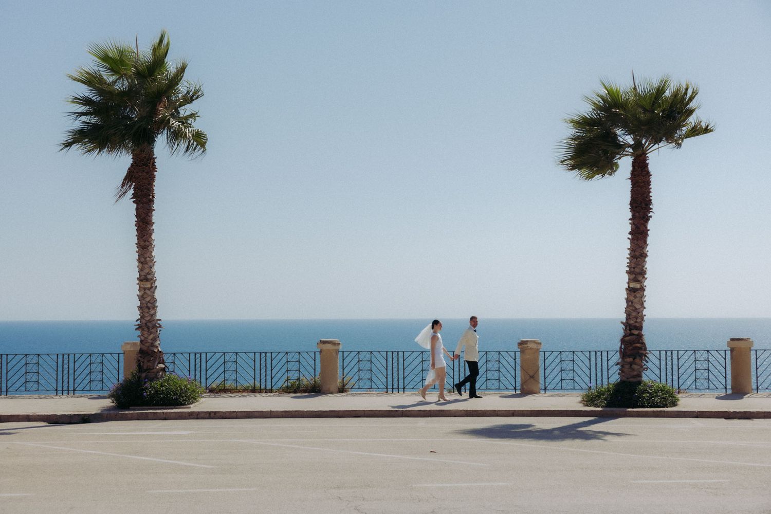 Palm trees line a beachfront walkway with blue railings overlooking the ocean on a sunny day.