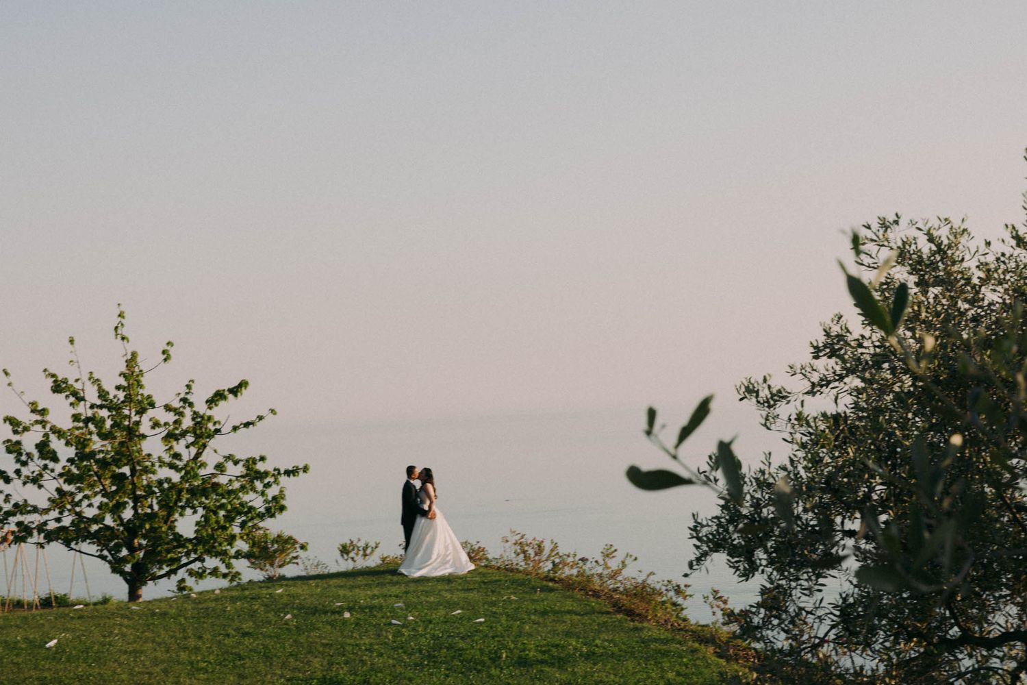 A couple embraces on a hilltop at sunset with trees silhouetted against the dusky sky.