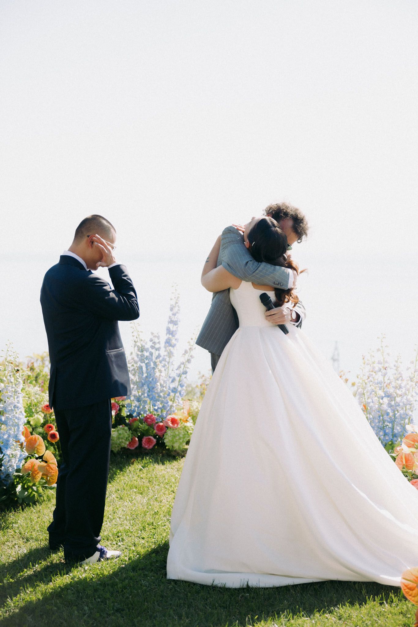 A romantic outdoor wedding ceremony with a couple embracing amid colorful flowers and blue delphiniums.