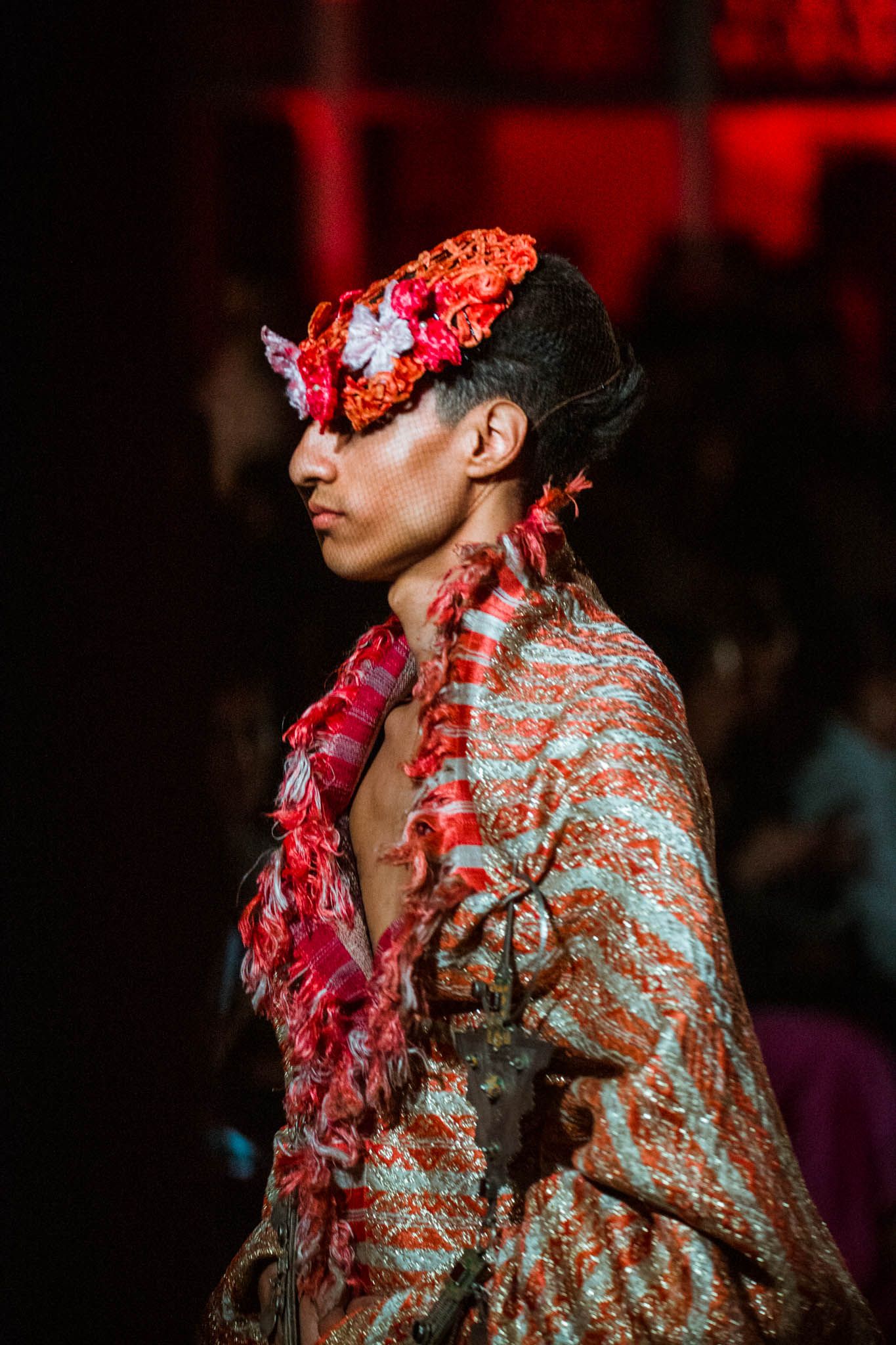Fashion model walks runway wearing ornate metallic jacket with dramatic red floral headpiece in dimly lit show.